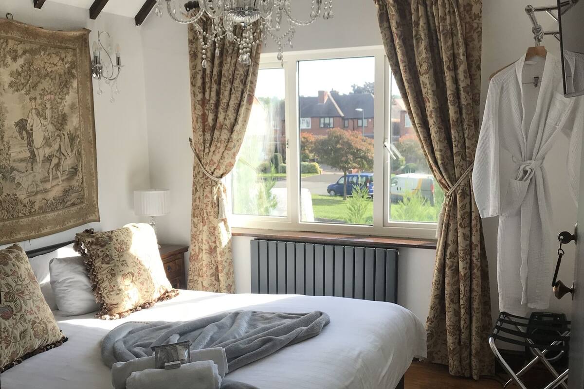 A bedroom with a large bed, a chandelier, a radiator, a window with curtains, and a view of the outside.