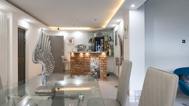 A modern dining area with a glass table and chairs, a decorative angel sculpture, a brick accent wall, and a bar area with shelves and bottles. - Achill Guest House (Solihull)