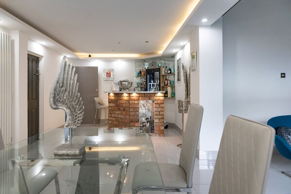 A modern dining area with a glass table and chairs, a decorative angel sculpture, a brick accent wall, and a bar area with shelves and bottles.