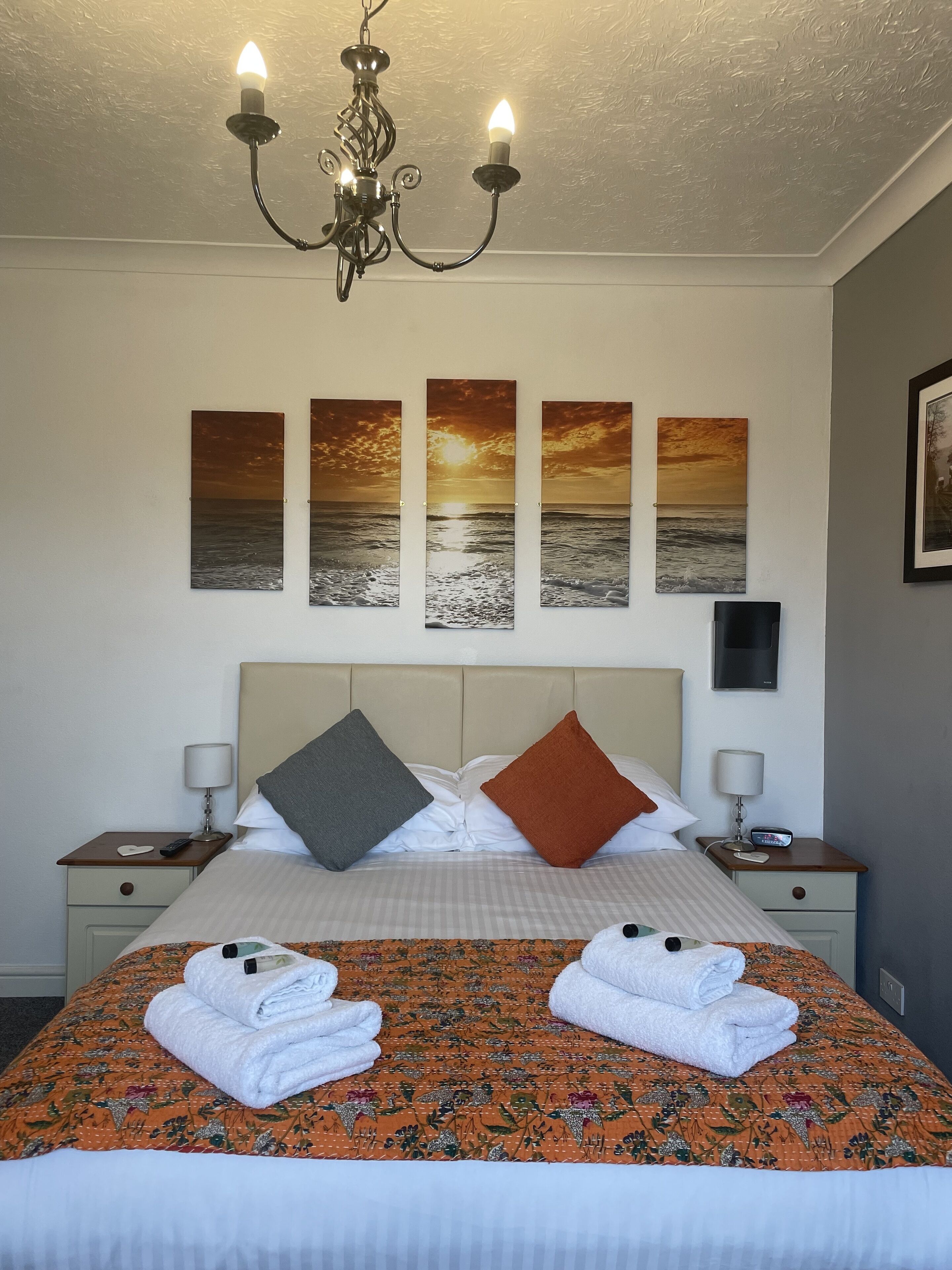 Une chambre d’hôtel avec un lit, deux tables de chevet, un lustre et trois tableaux représentant des couchers de soleil accrochés au mur.