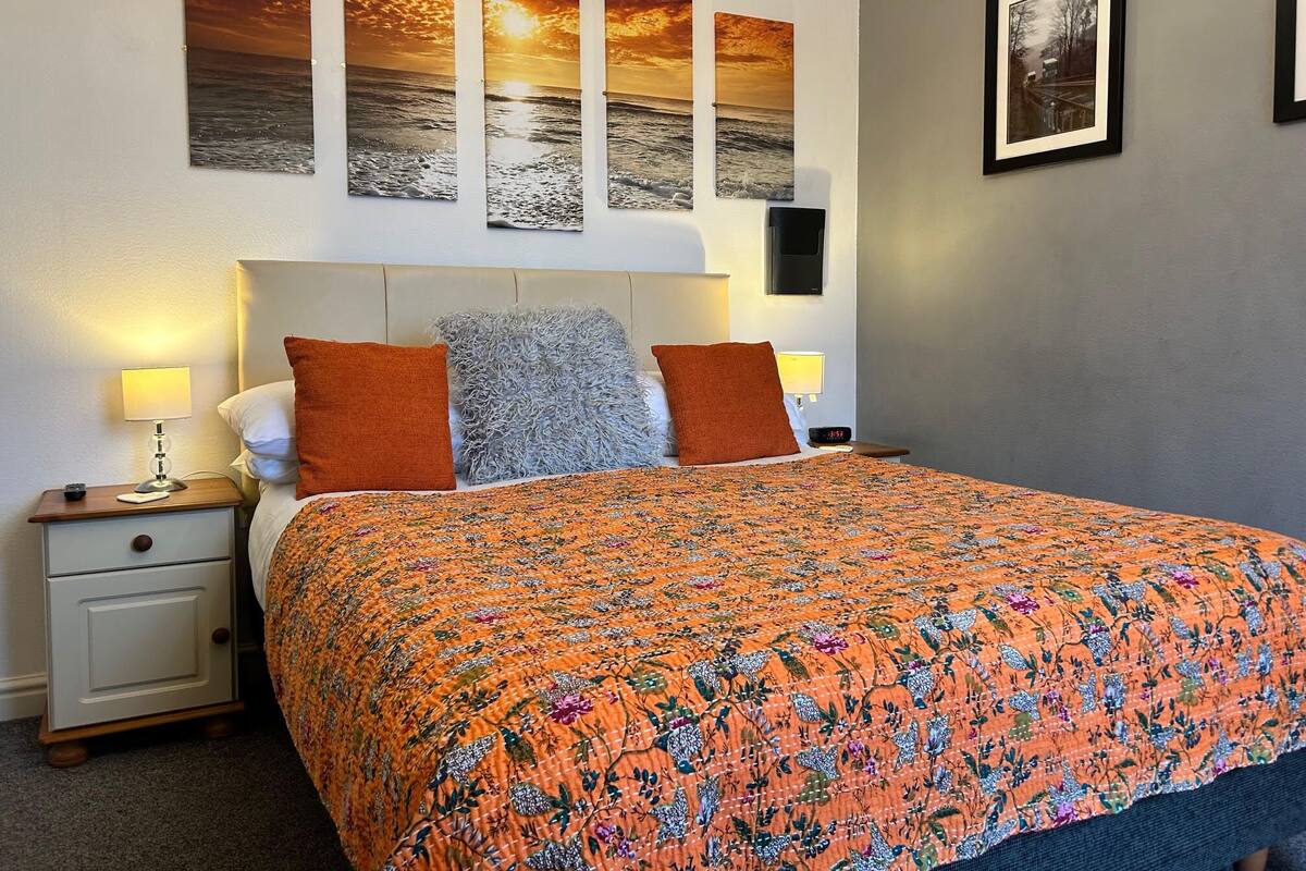 Een opgemaakt bed met een oranje bloemenbedekking, twee kussens en een nachtkastje met een lamp.