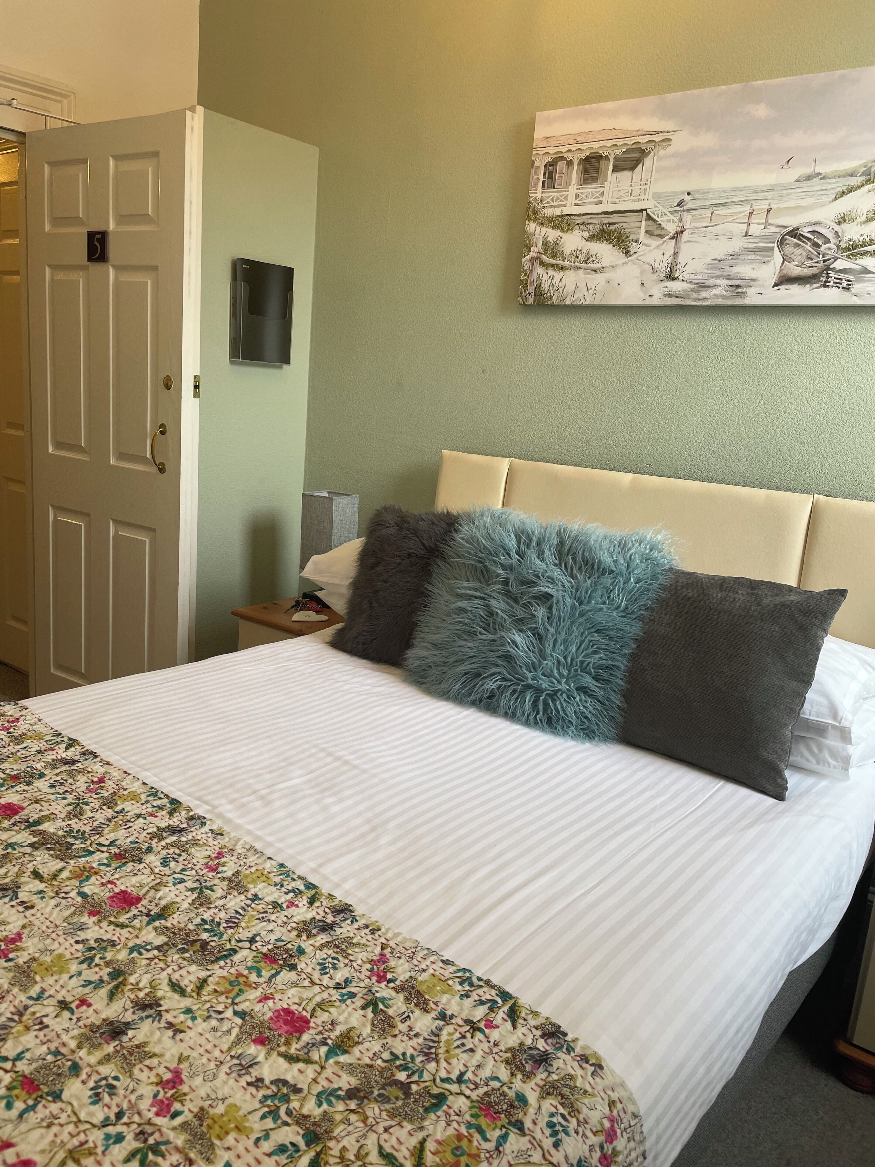 Una cama bien hecha con una colcha de estampado floral y dos almohadas, una obra de arte colgada en la pared que representa una escena costera, y una puerta con un número.