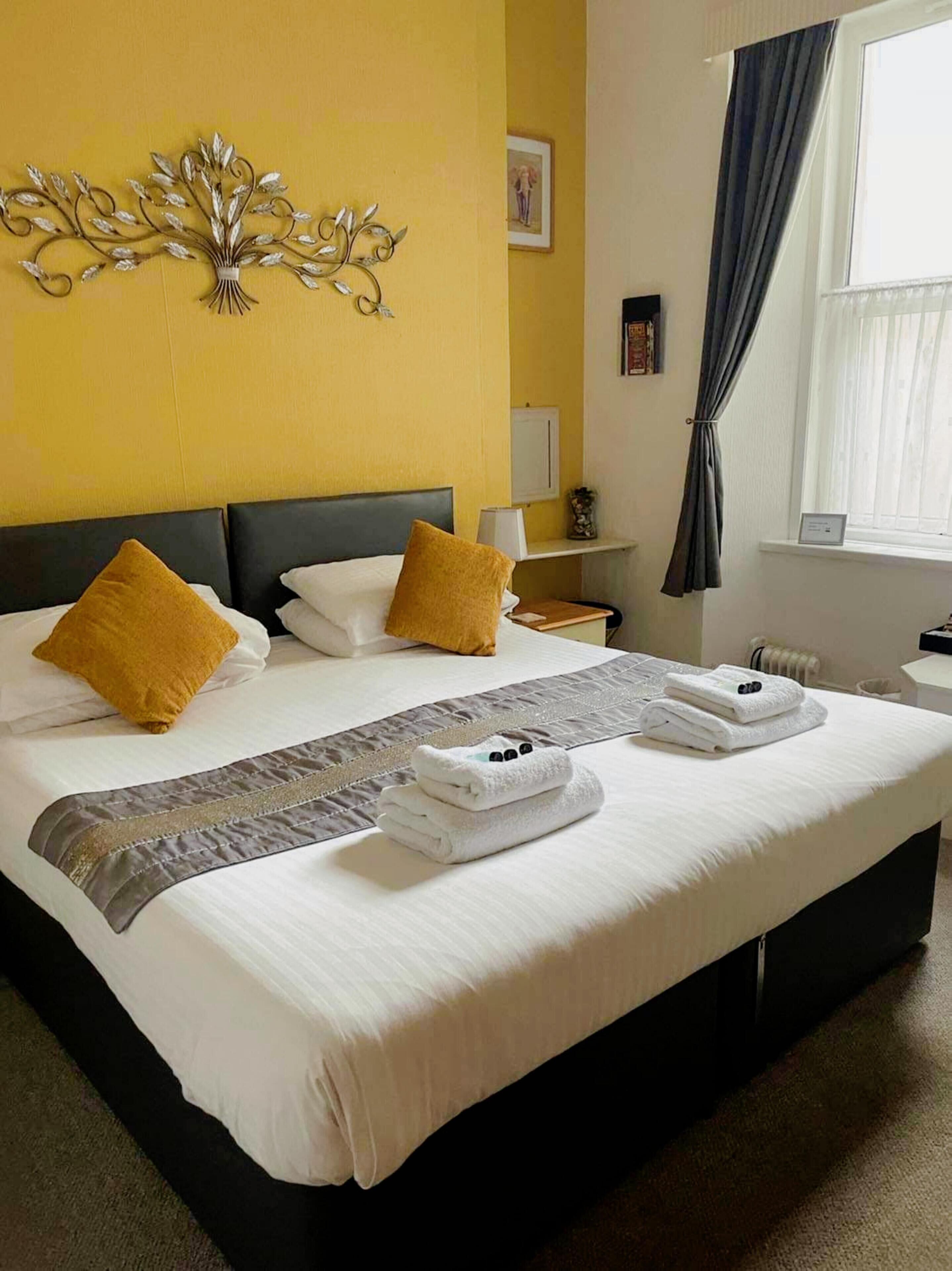 Quarto de hotel com uma cama grande, duas almofadas, um ornamento na parede, uma mesa de cabeceira com abajur e uma janela com cortinas.