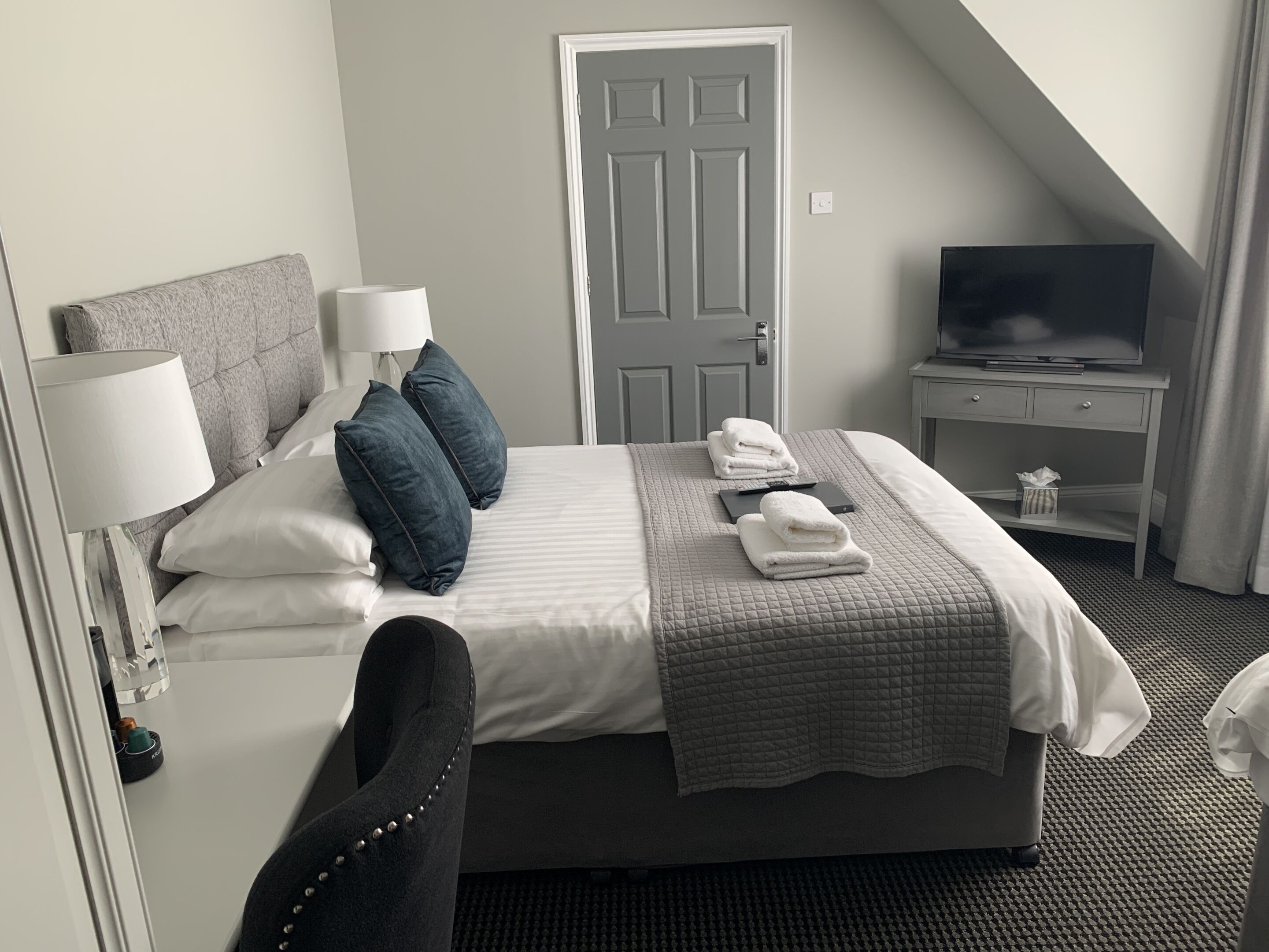 A hotel room with a bed, pillows, a TV, a bedside table, and a chair.