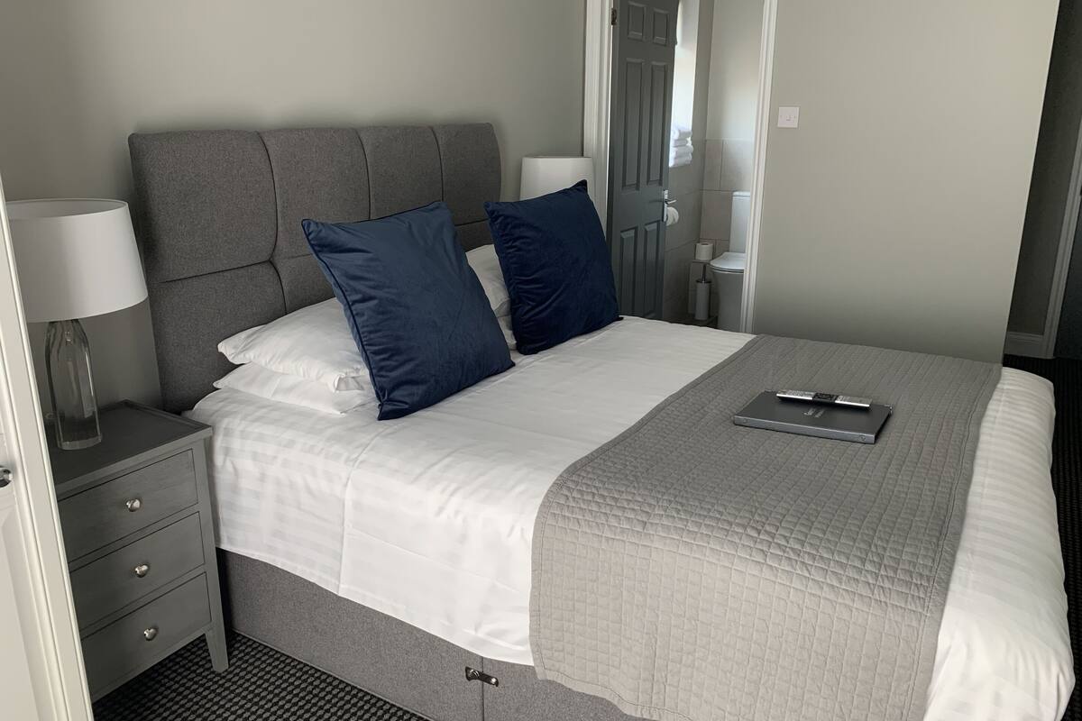 Una cama bien tendida con un cabecero gris, dos almohadas azul oscuro y una colcha gris acolchada. Hay una mesita de noche con una lámpara y un control remoto encima.