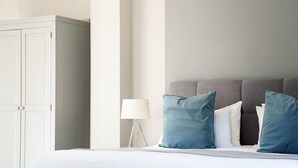 Una cama bien hecha con almohadas blancas y azules, cabecera gris y mesita de noche blanca con una lámpara.
