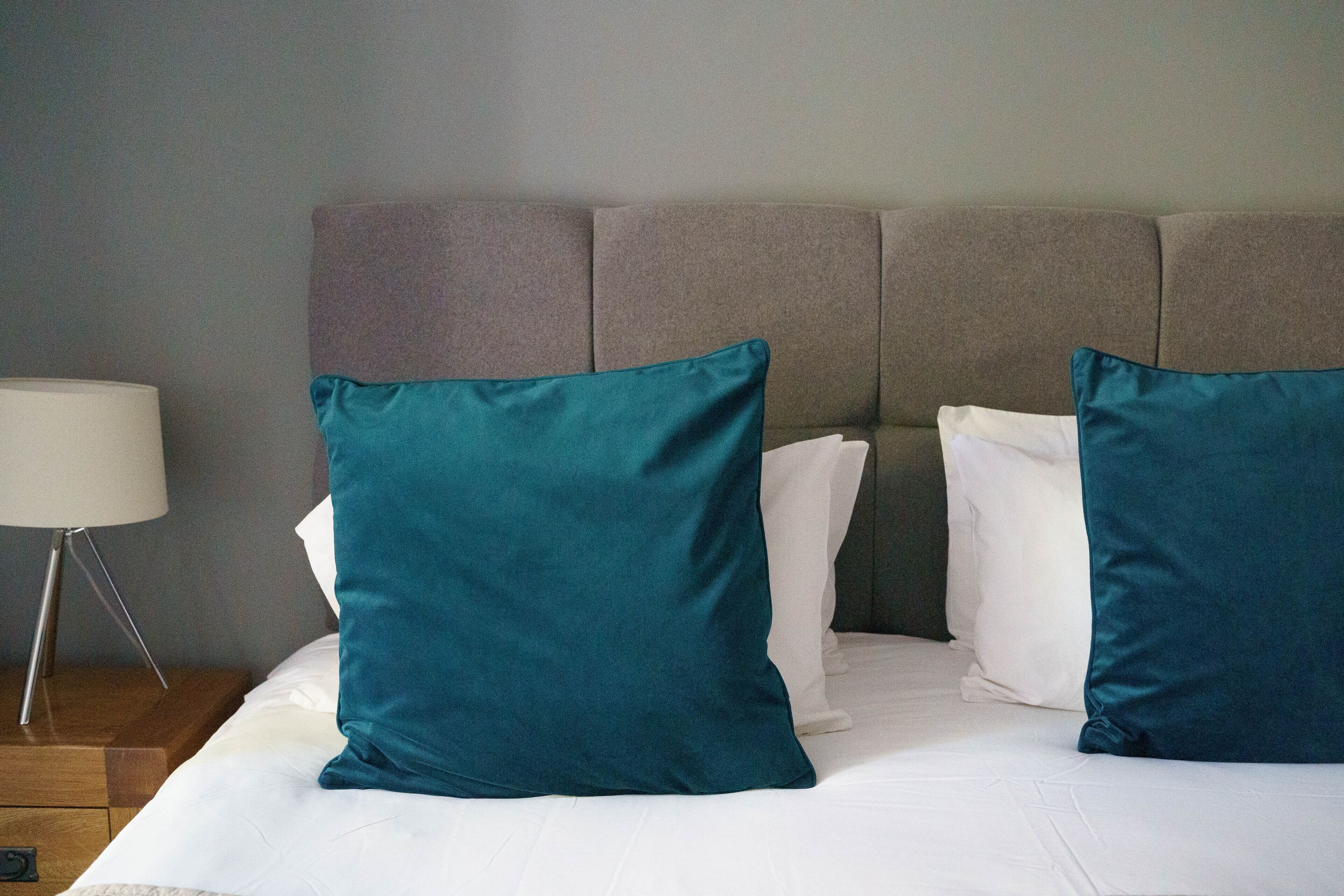 Una cama bien hecha con sábanas blancas y almohadas turquesa, una lámpara de noche y una mesita de madera.