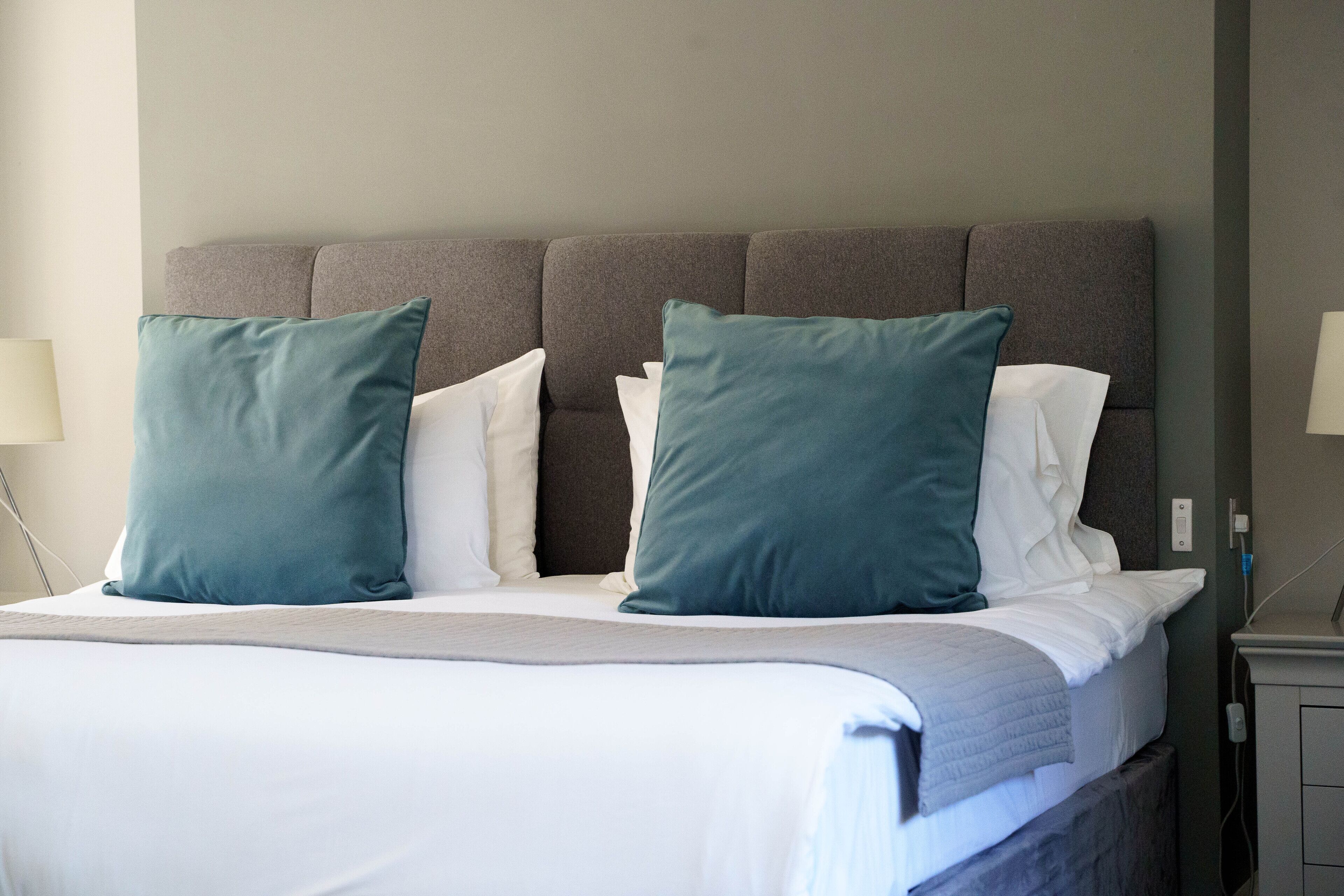 Una cama bien hecha, con un cabecero gris, almohadas blancas y azul verdosas, y una colcha gris.
