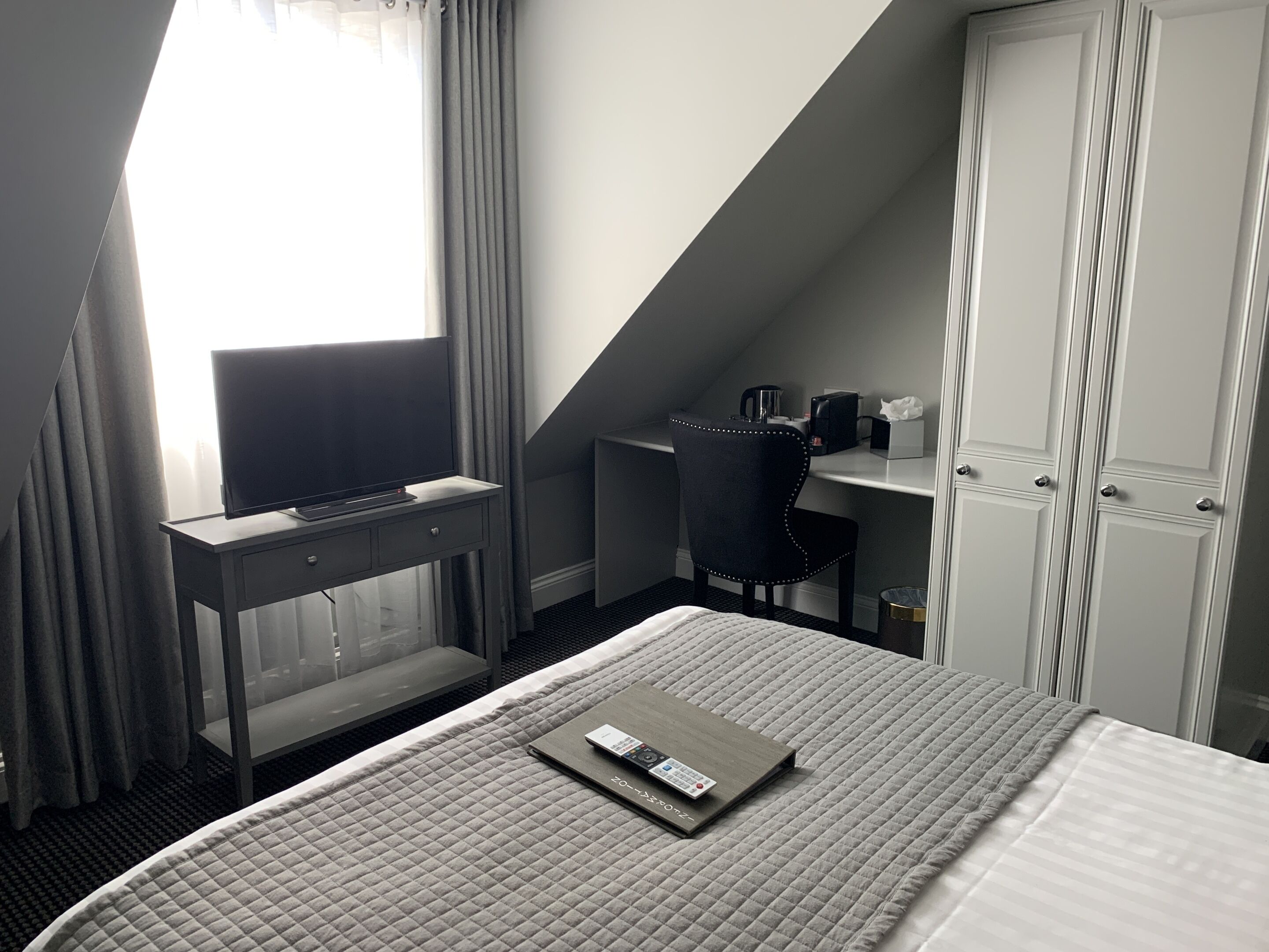 A hotel room with a bed, a desk with a TV, a chair, a wardrobe, and a window with curtains.