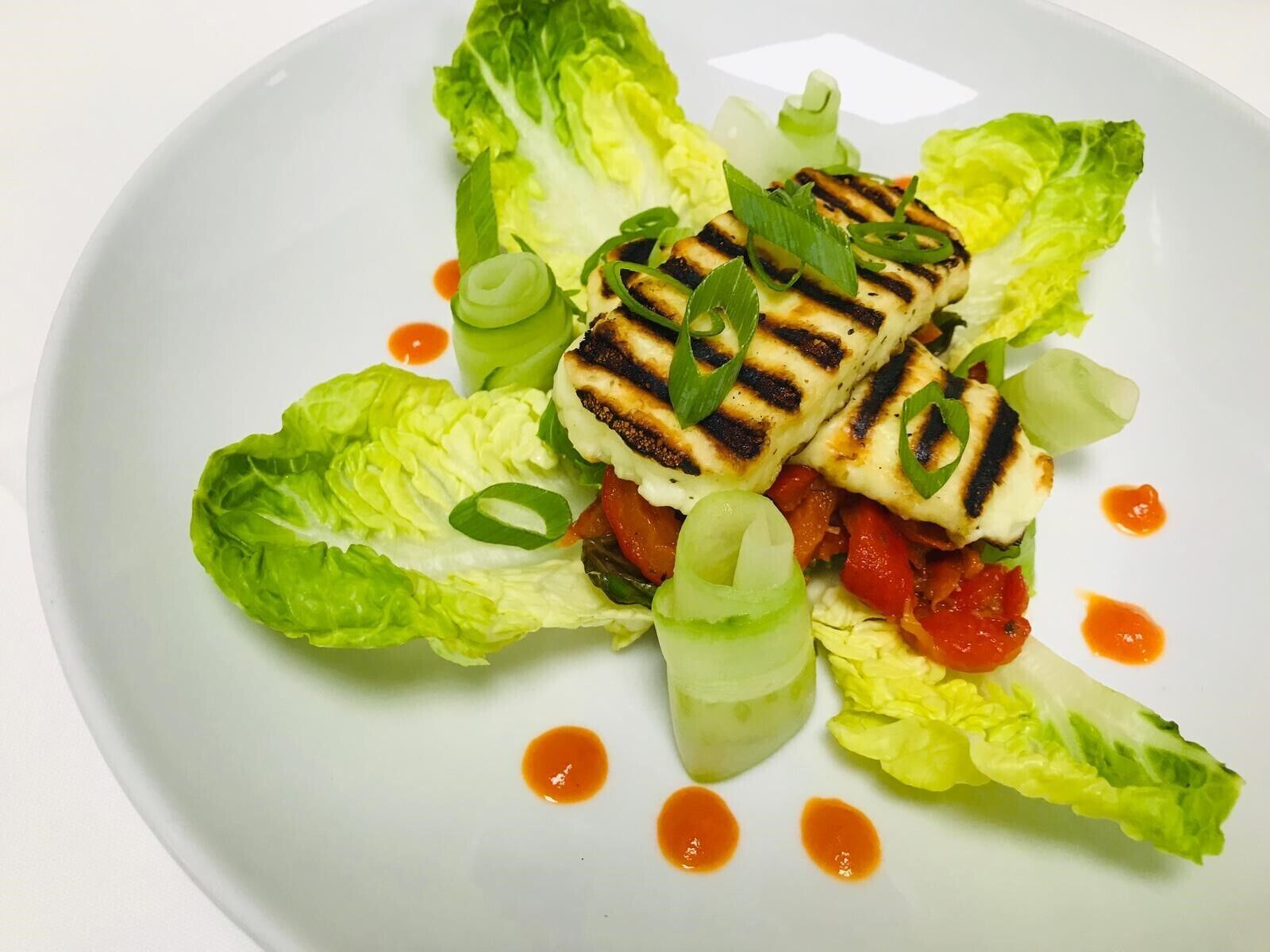 Een bord met gegrilde kip, sla en plakjes tomaat, versierd met groene uien en overgoten met saus.