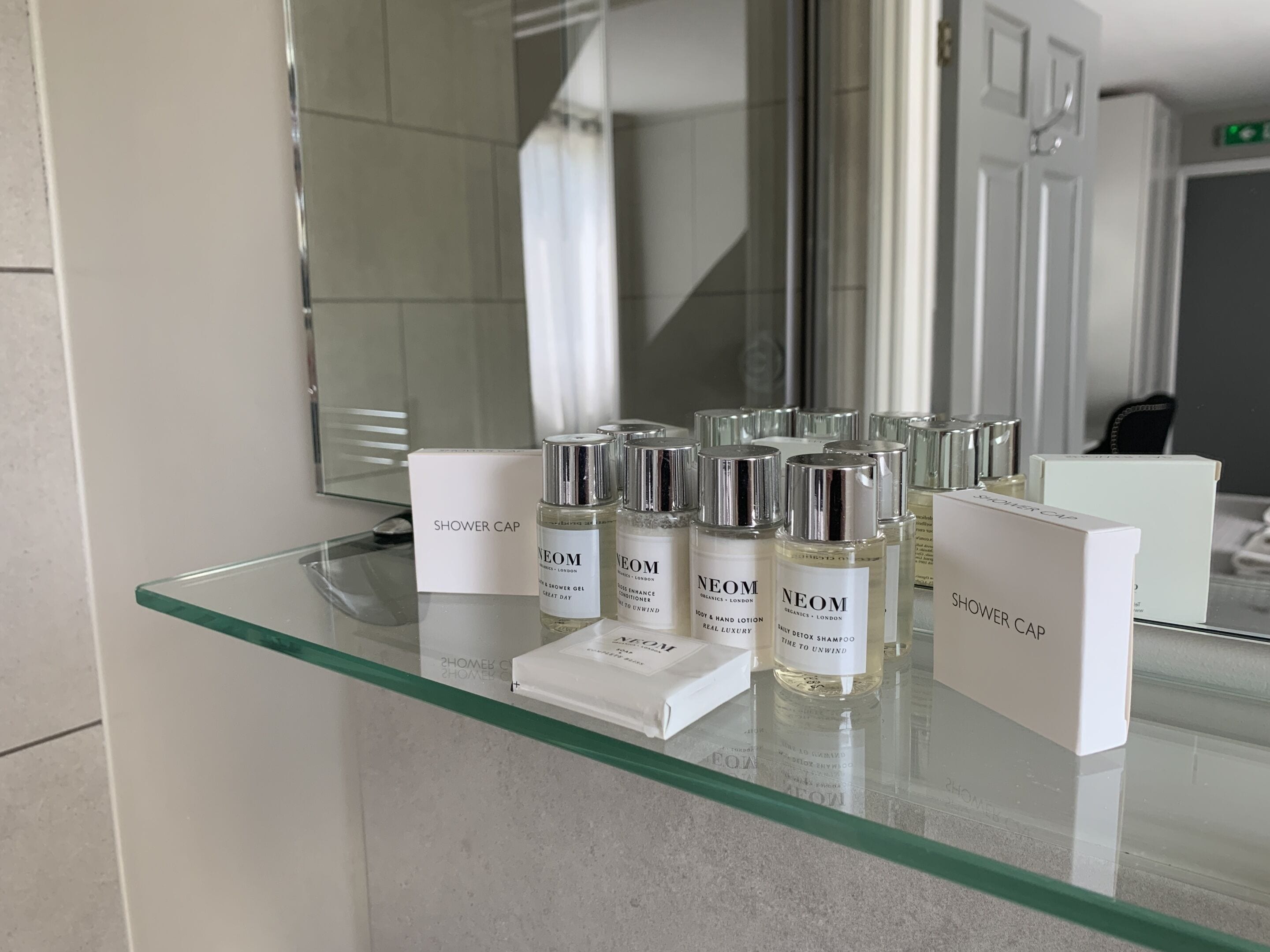 A bathroom with a glass shelf displaying various Neom products, including shower caps and bottles of lotion and shampoo.