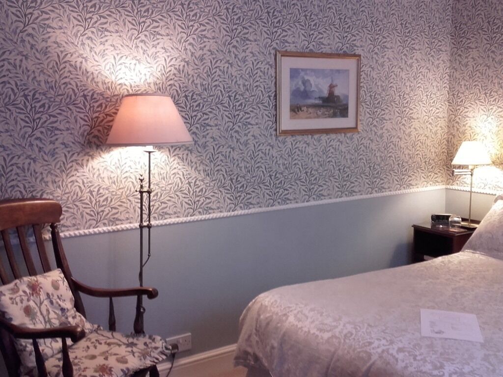 A bedroom with a patterned wallpaper, a framed picture on the wall, a wooden chair, and two bedside lamps.