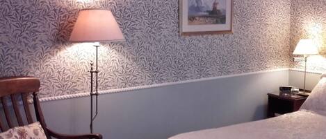 A bedroom with a patterned wallpaper, a framed picture on the wall, a wooden chair, and two bedside lamps.