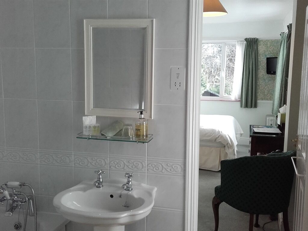 A bathroom with a white sink, mirror, and glass shelves. There is a shower area with a handheld showerhead and a towel rack. The room has a window with curtains and a bed in the background.