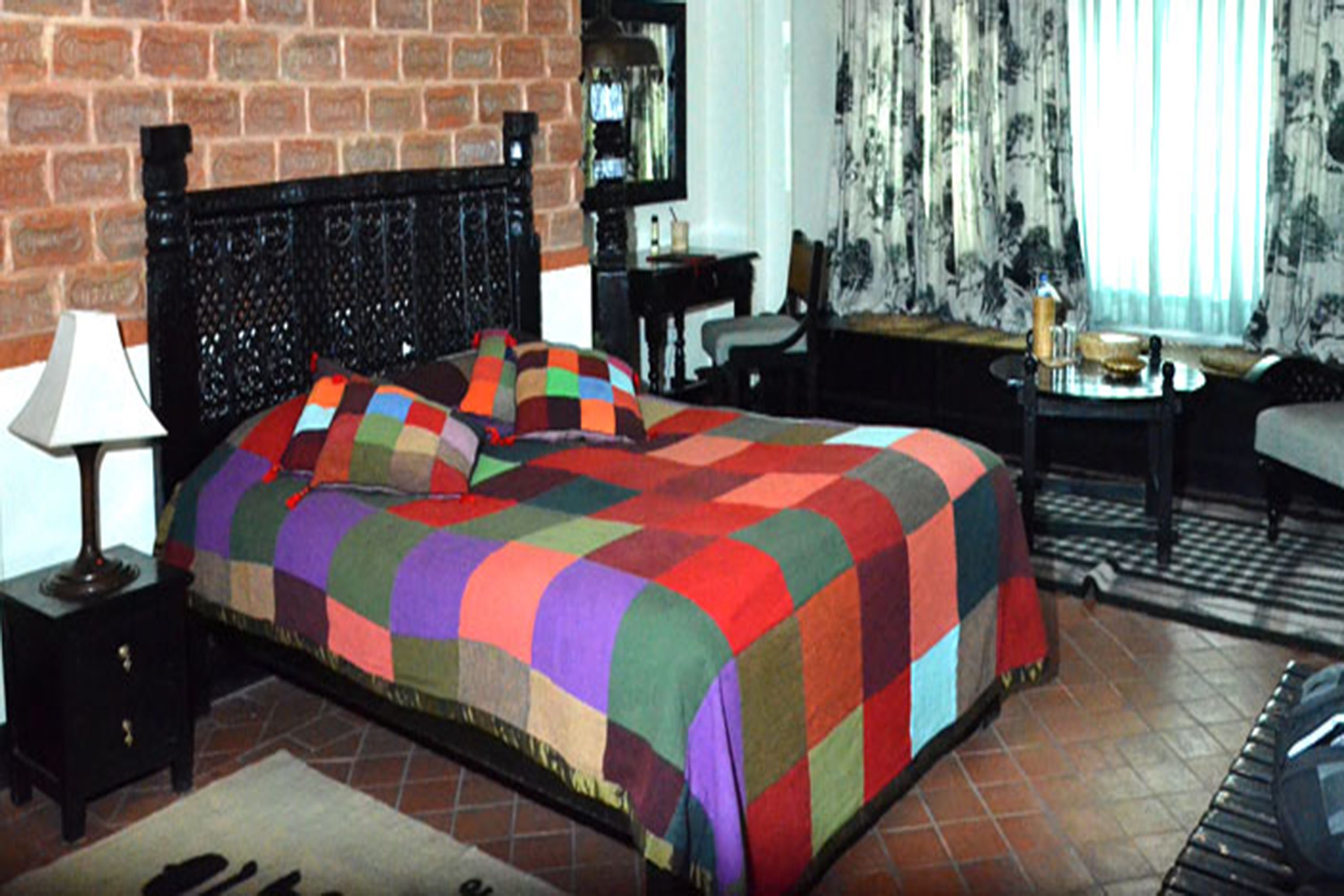 Une chambre avec un mur en briques, une tête de lit noire, une courtepointe colorée, une lampe, une chaise, une table et une fenêtre avec des rideaux.