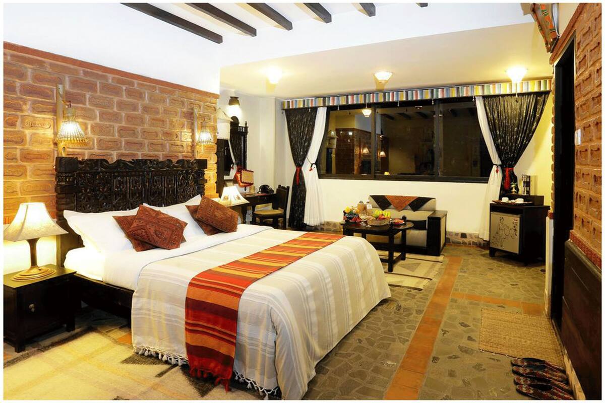 A hotel room with a brick wall, a large bed with a striped blanket, a nightstand with a lamp, a small table with a lamp, and a window with curtains.