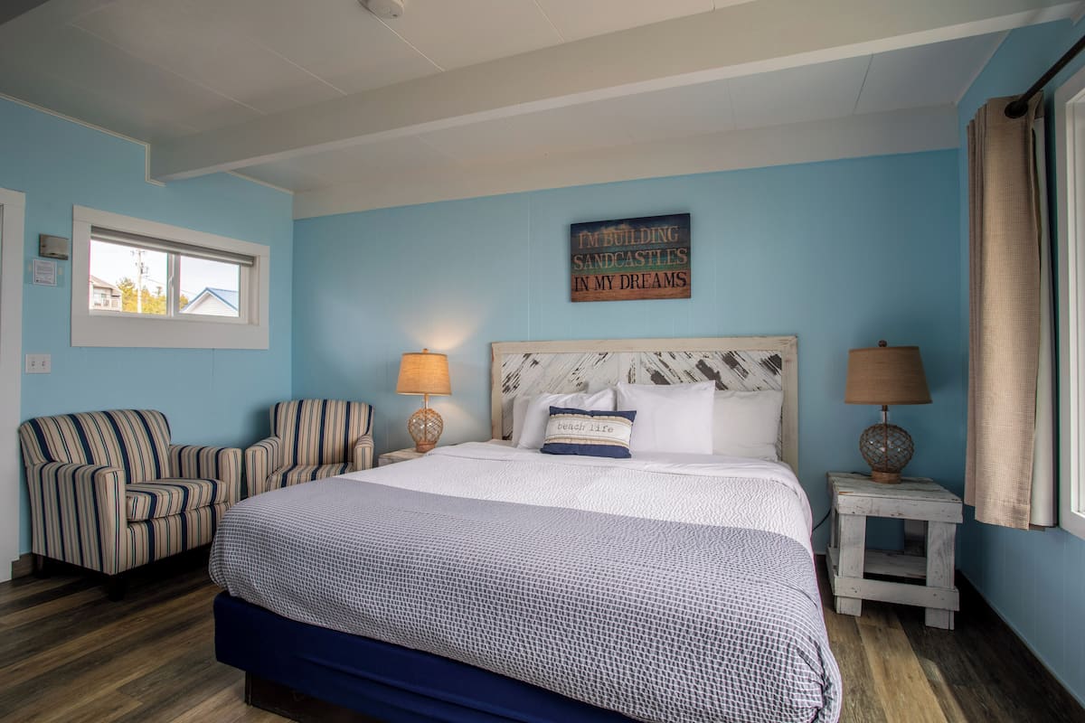 A bedroom with a large bed, two bedside tables with lamps, a striped armchair, a window, and a wall sign.