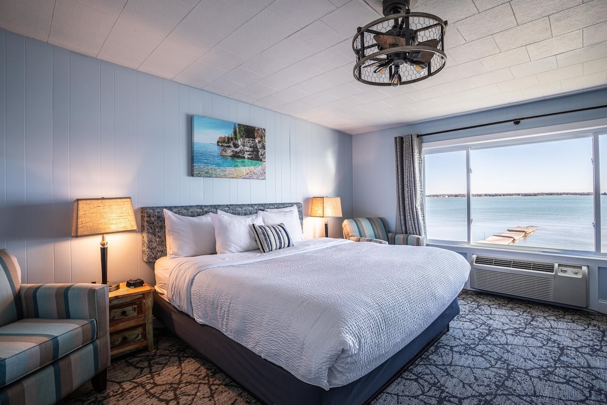 A hotel room with a large bed, two bedside lamps, a chair, a painting of a coastal scene, and a view of the sea through large windows.