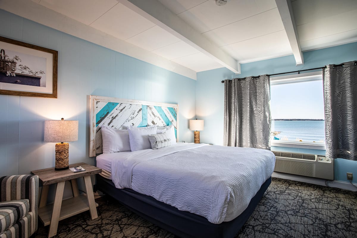 A hotel room with a large bed, two bedside lamps, a view of the sea through a window, and a framed picture on the wall.