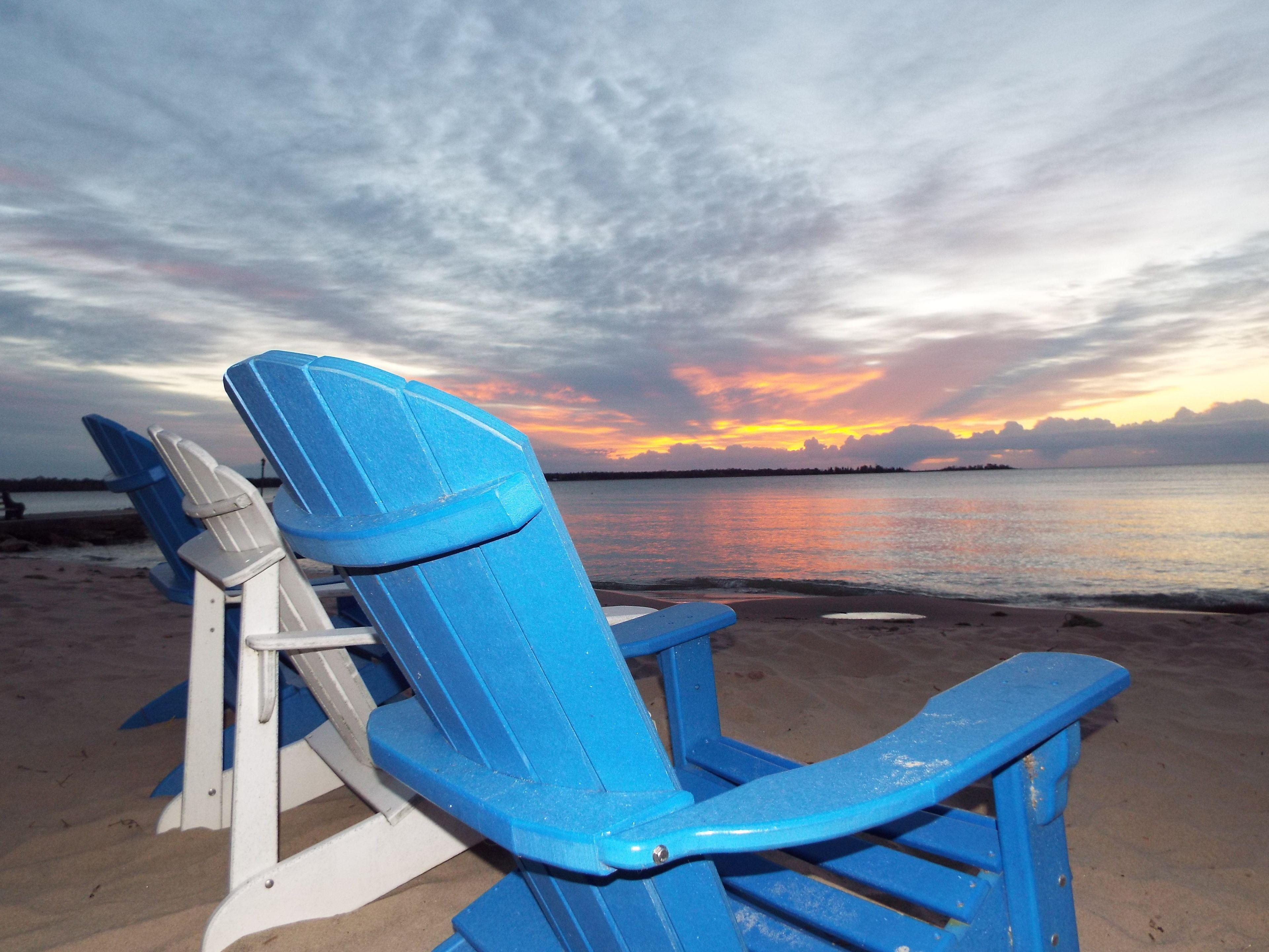 A beach with blue Adirondack chairs and a sunset over the water.
