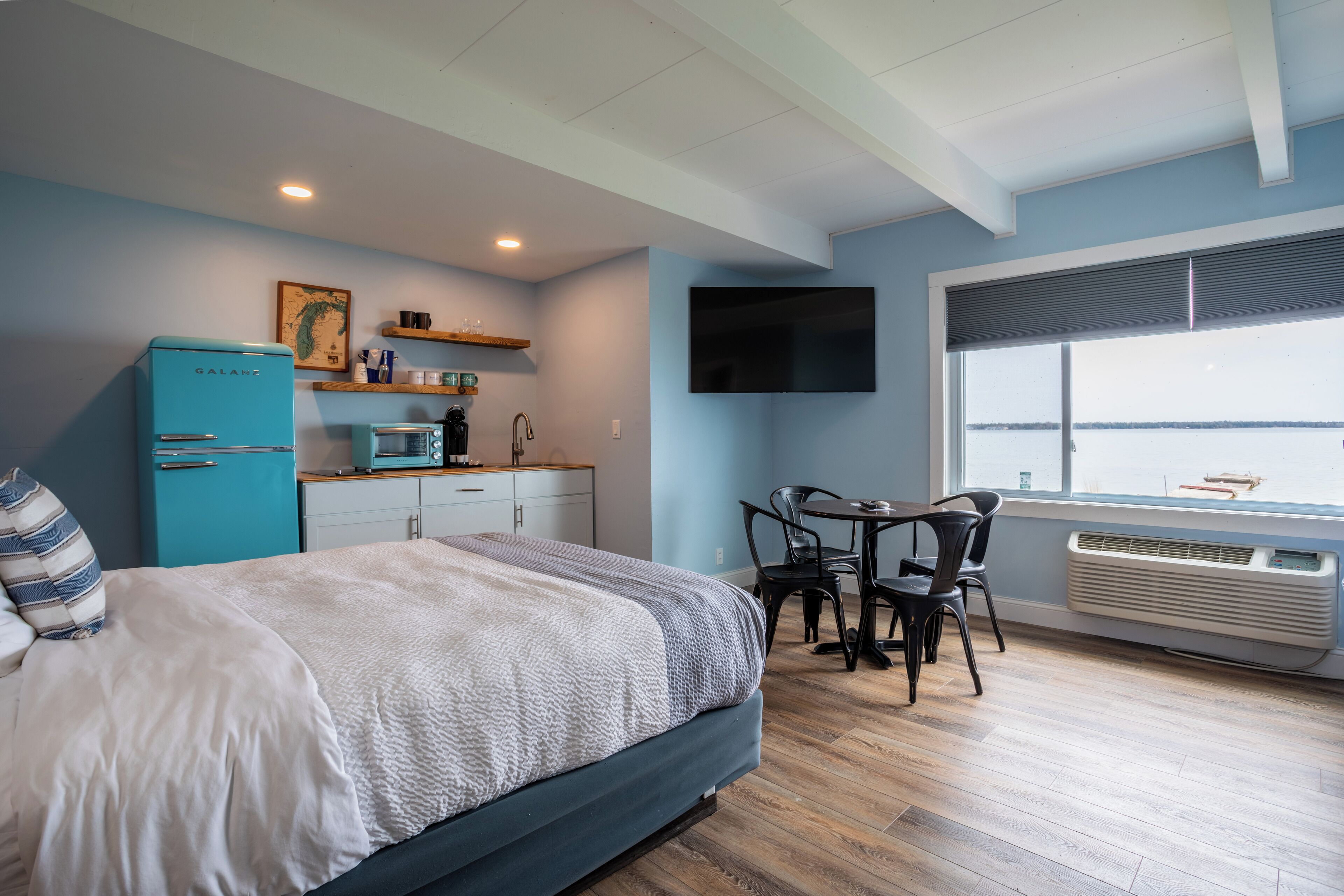 A bedroom with a bed, a desk, a chair, a blue refrigerator, a microwave, a TV, and a window with a view.