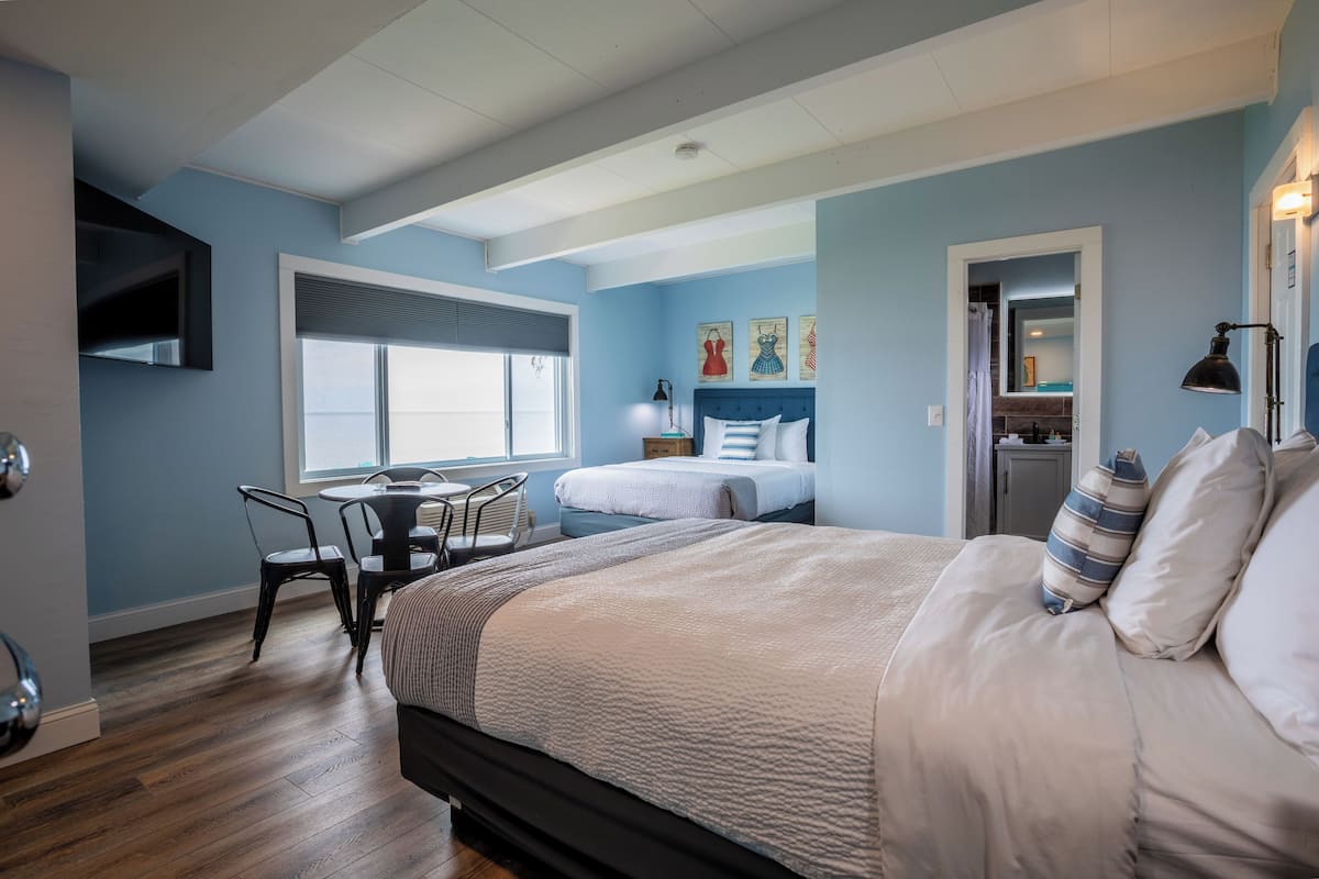 A bedroom with a large bed, a desk, and a chair. There is a window with blinds, a door, and a small cabinet.