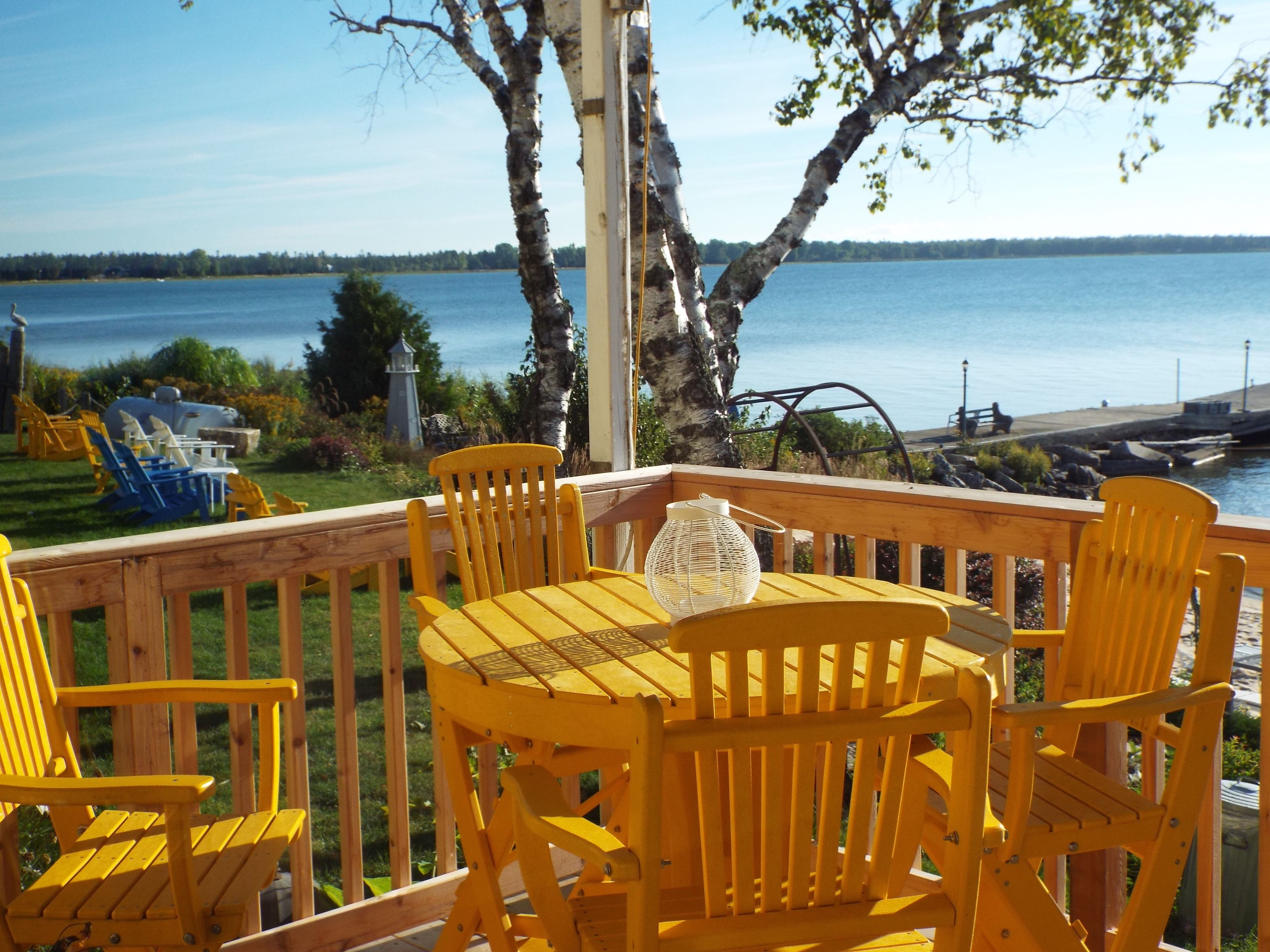 Une terrasse en bois avec des chaises jaunes et une table, d’où l’on peut admirer un plan d’eau avec un phare au loin.