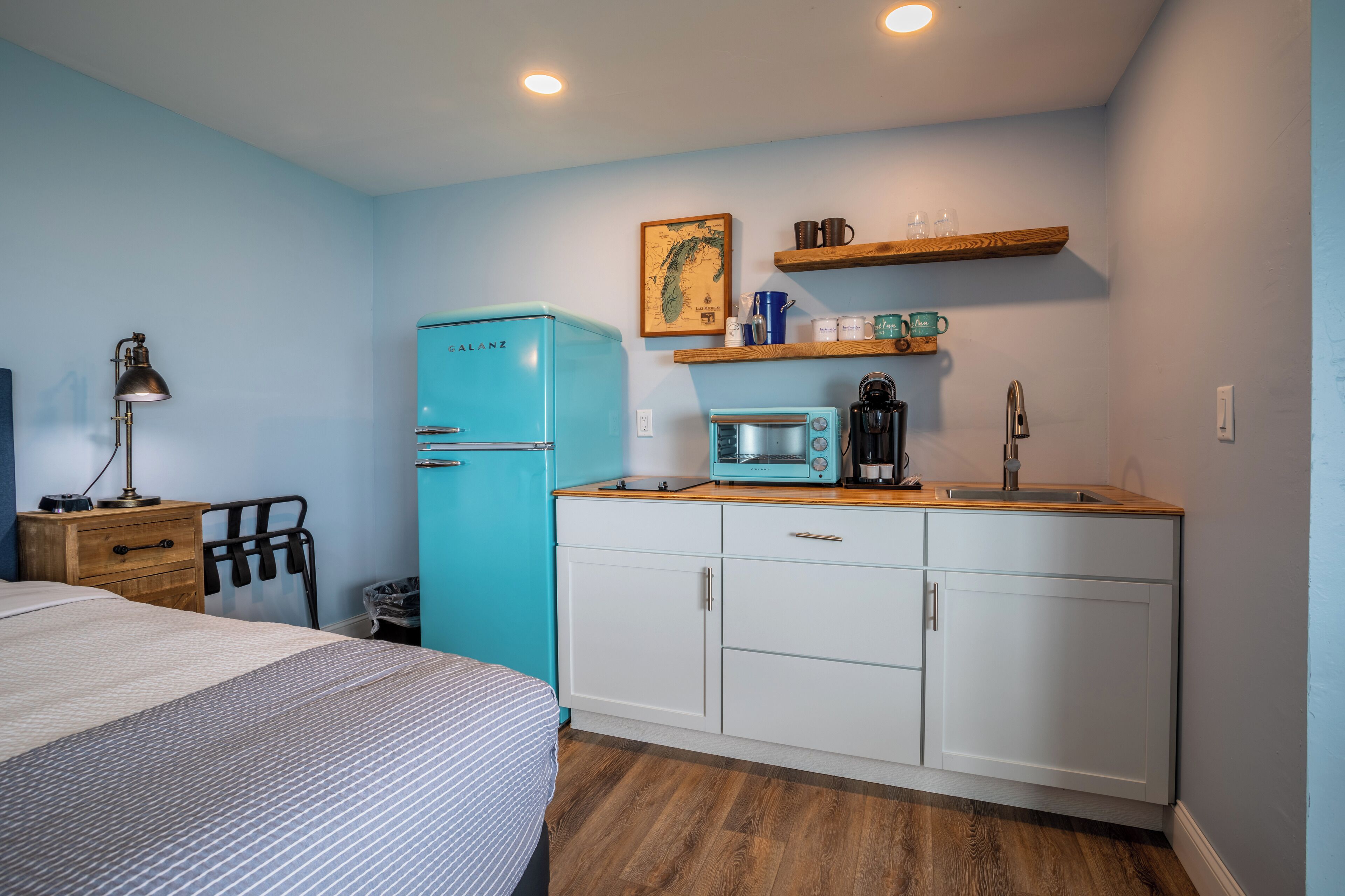 Habitación compacta con cama, una kitchenette pequeña con microondas, refrigerador y estante de madera sobre el lavamanos.
