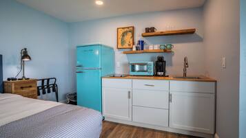 Habitación compacta con cama, una kitchenette pequeña con microondas, refrigerador y estante de madera sobre el lavamanos.
