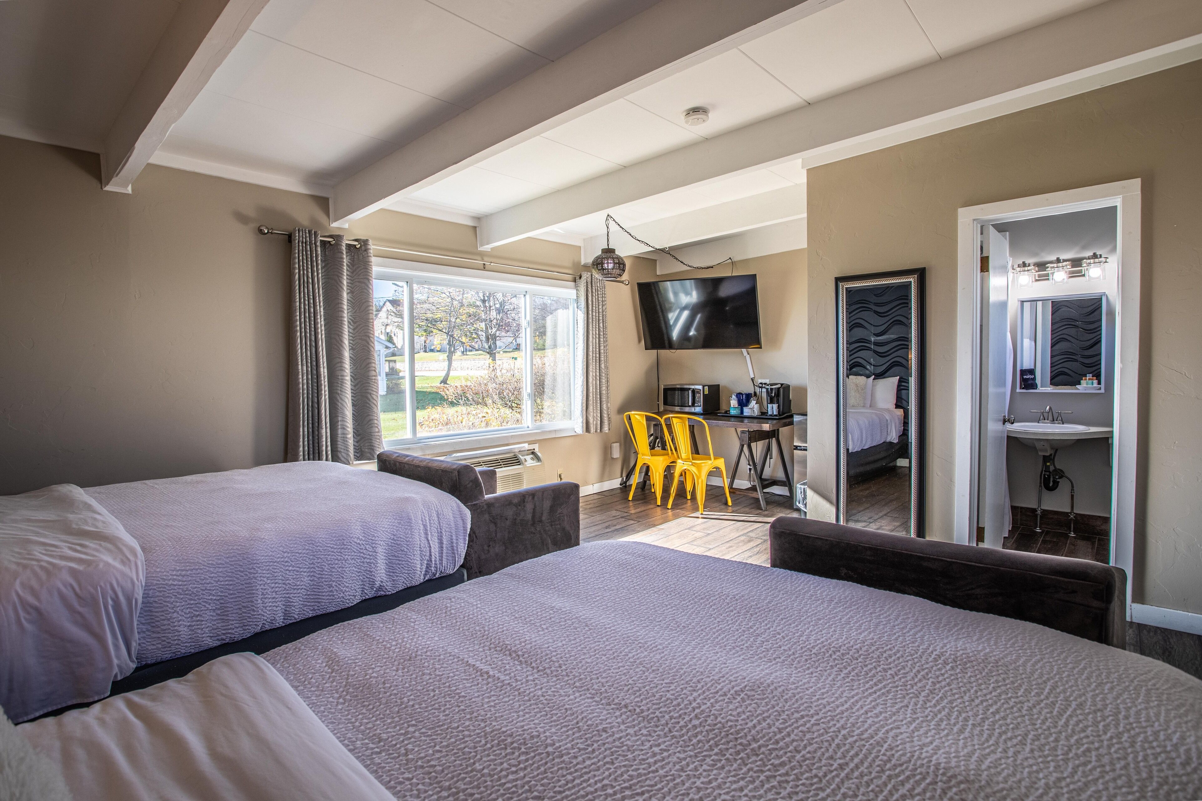 A hotel room with two beds, a TV, a desk with a chair, and a bathroom with a mirror and sink.