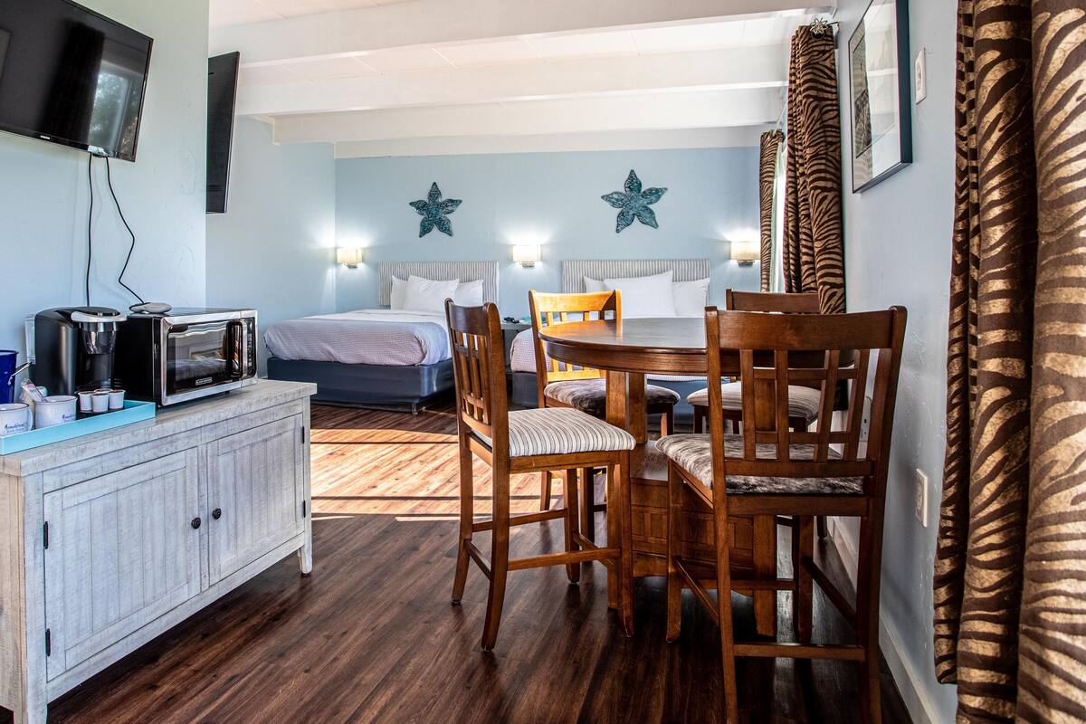 A hotel room with a bed, a dining table with chairs, a TV mounted on the wall, and a kitchenette area.