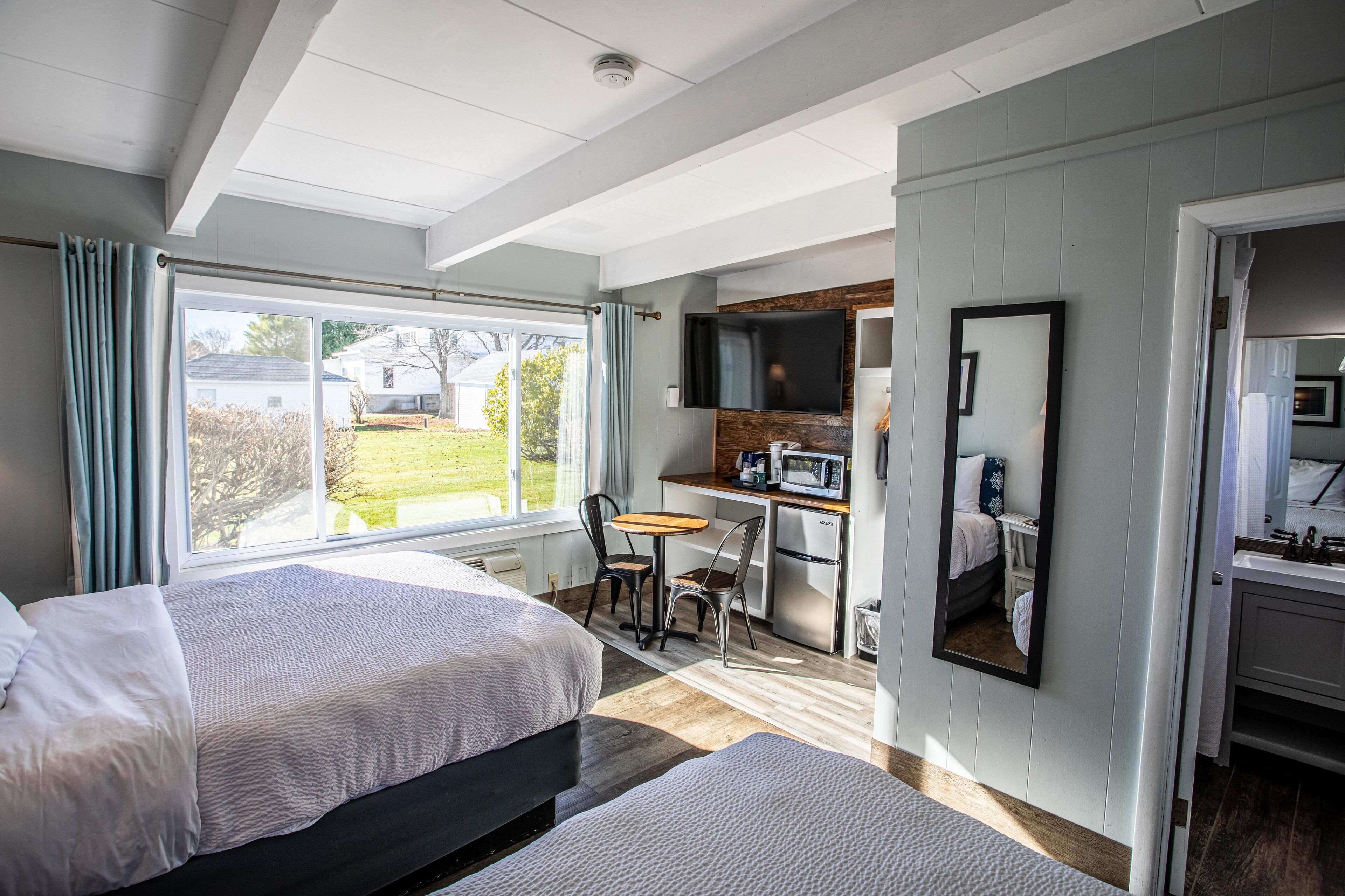A hotel room with two beds, a kitchenette, a TV, and a large window offering a view of the outdoors.