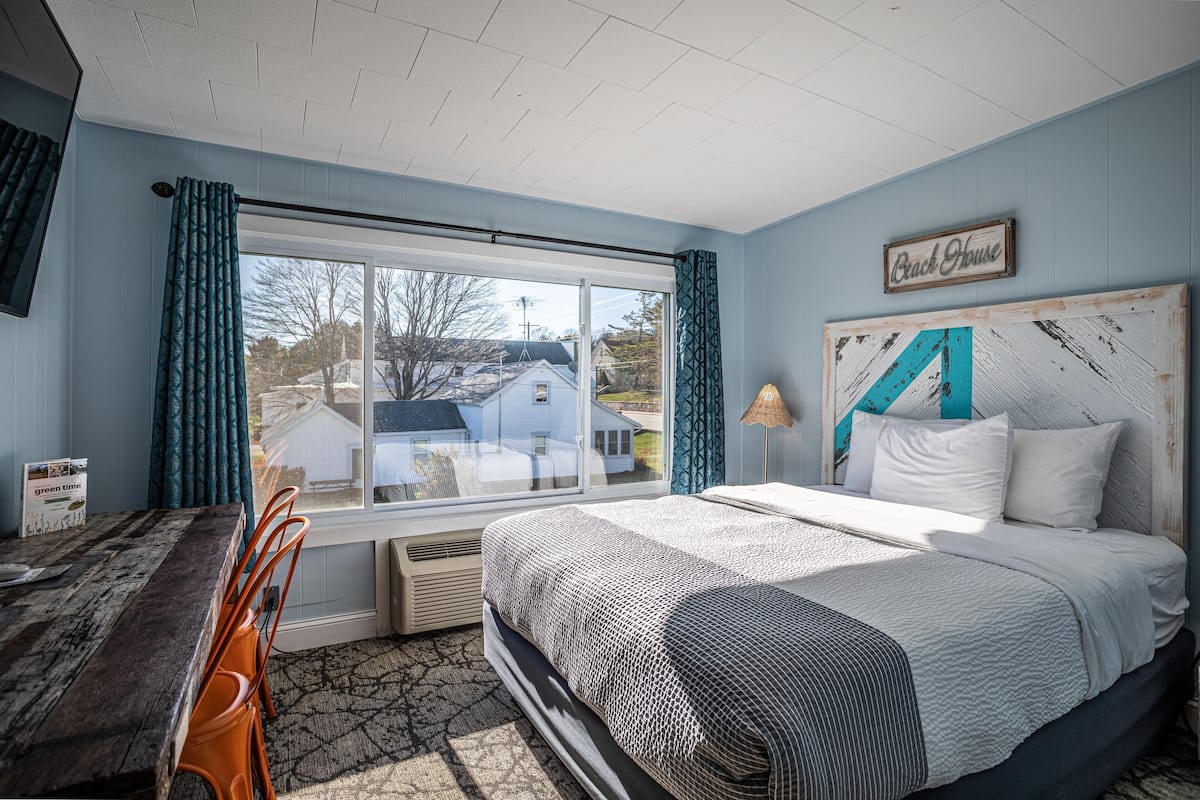 A bedroom with a large window, a bed with a white and blue headboard, and a view of a house outside.
