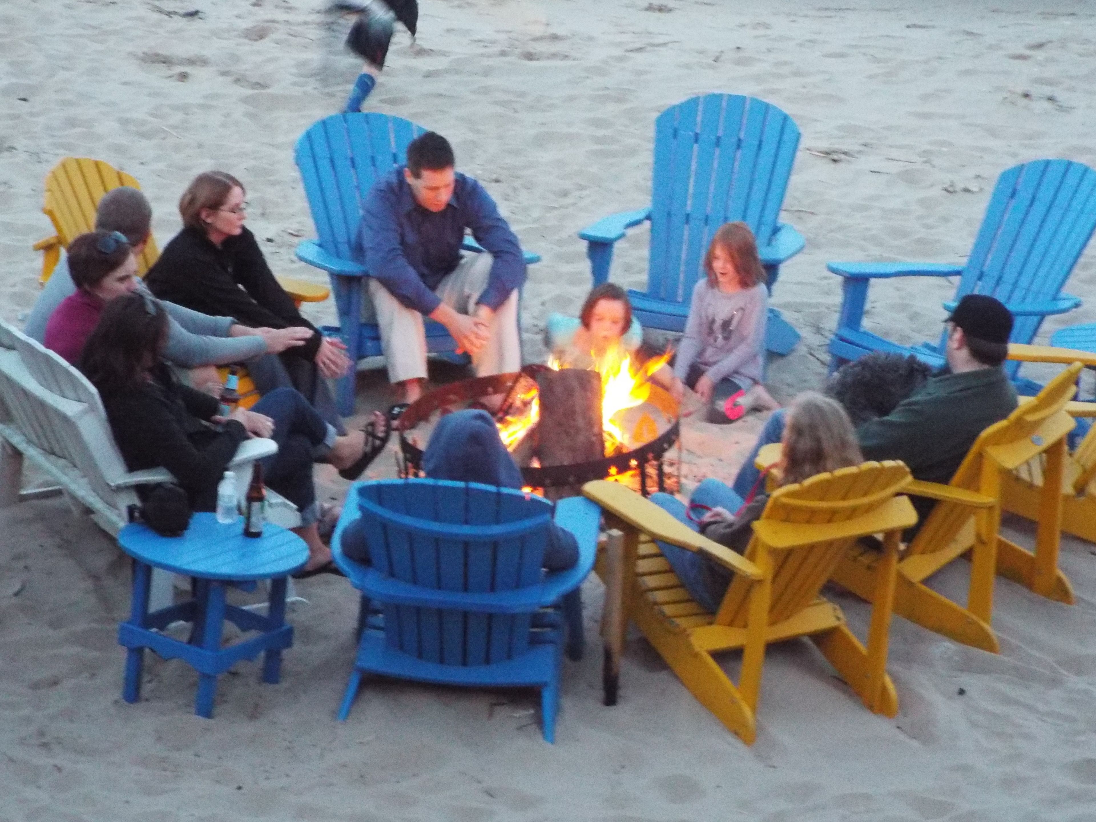A group of people seated around a campfire on a sandy beach with blue and yellow Adirondack chairs.
