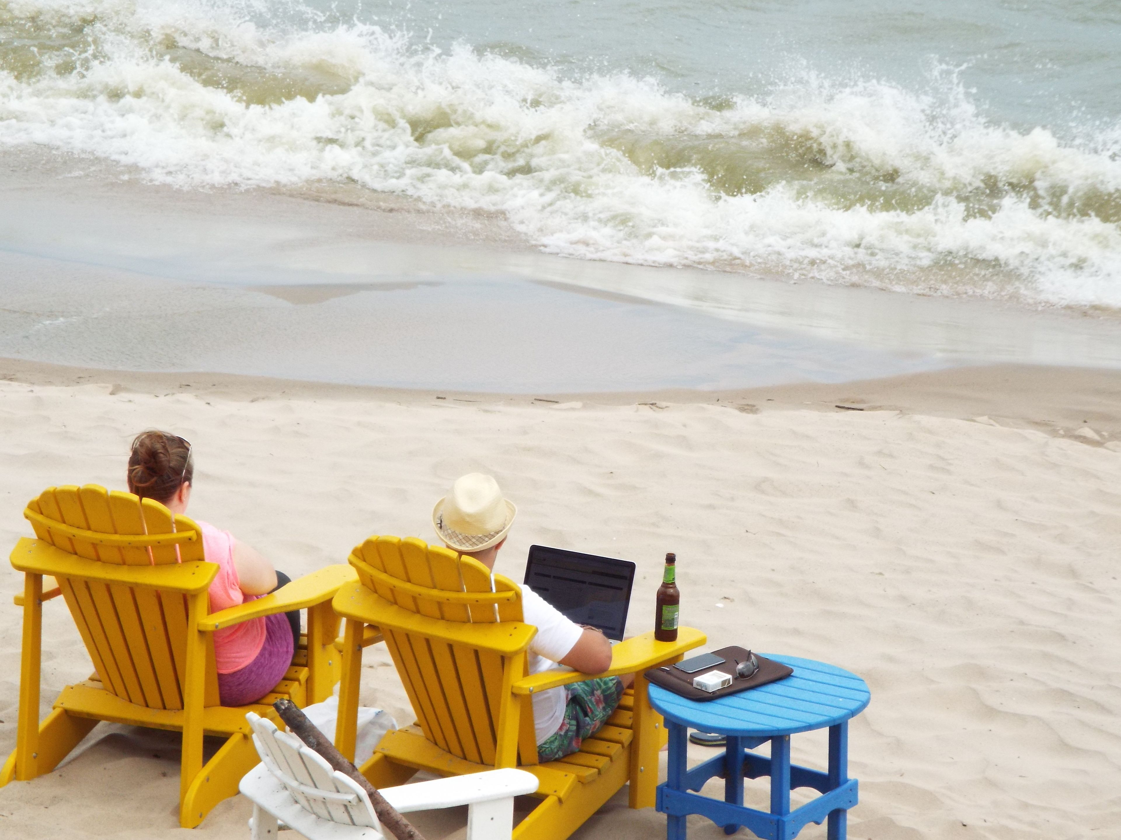 Twee personen zitten in gele Adirondack stoelen op een zanderig strand, met een laptop en een fles op een klein blauw tafeltje.