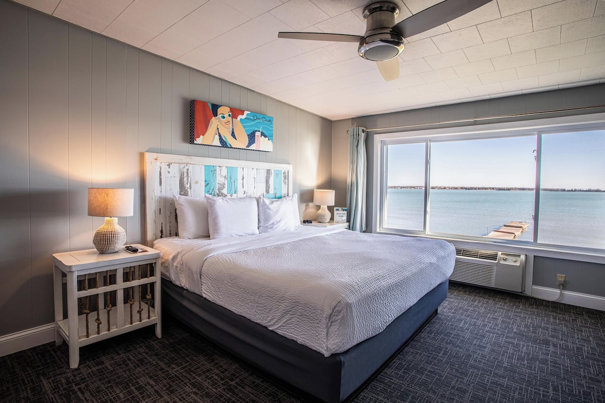 A hotel room with a large bed, a ceiling fan, a window with a view of the water, and a painting on the wall.