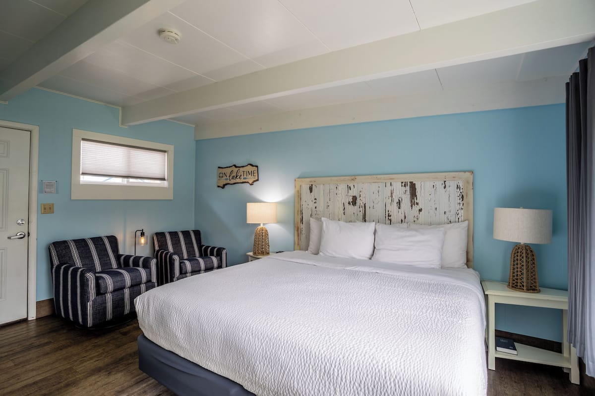A bedroom with a large bed, two armchairs, a nightstand with lamps, a window with blinds, and a sign on the wall.