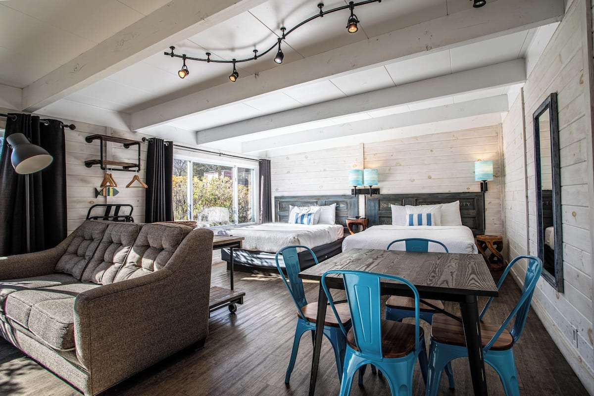 A modern hotel room with a sofa, a dining table with chairs, two beds, and a large window.