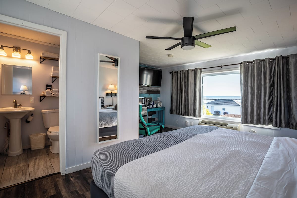 A bedroom with a bed, a desk, a chair, a window with curtains, a ceiling fan, and a view of the outside.