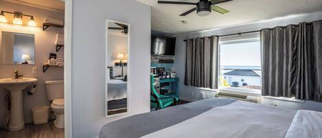 A bedroom with a bed, a desk, a chair, a window with curtains, a ceiling fan, and a view of the outside.