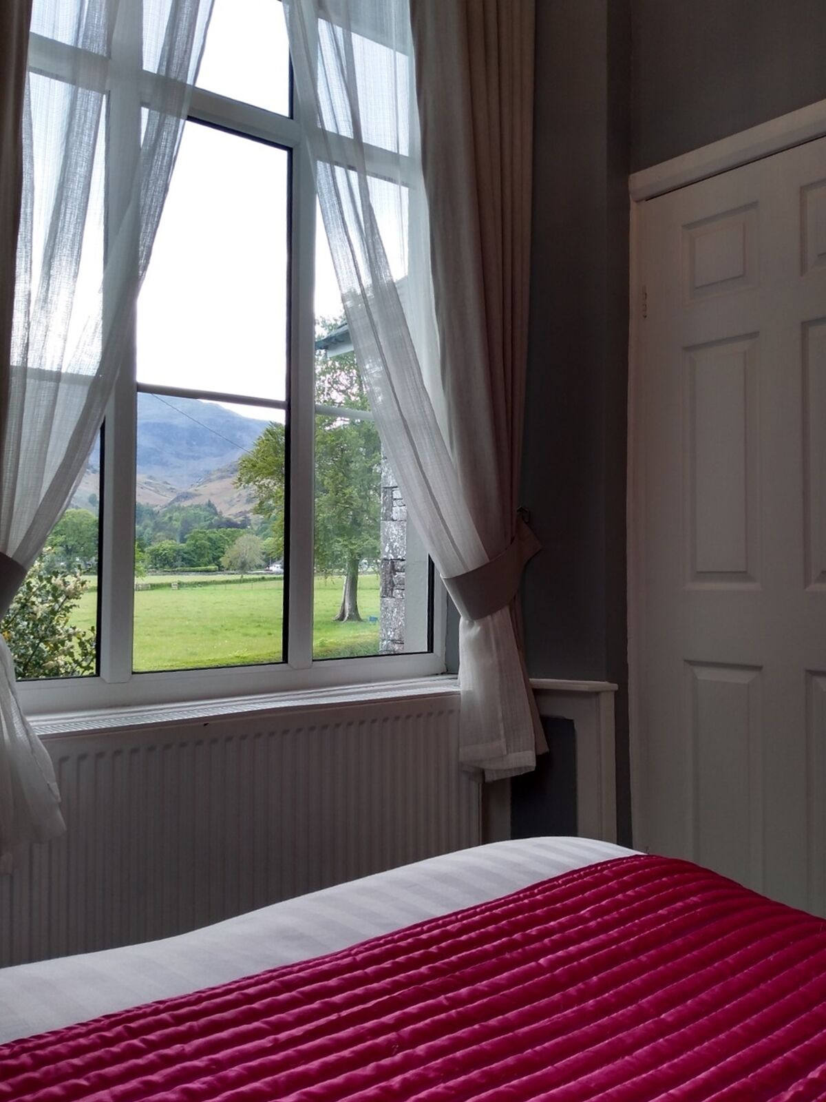 A bedroom with a bed, a window with sheer curtains, and a view of a green landscape.