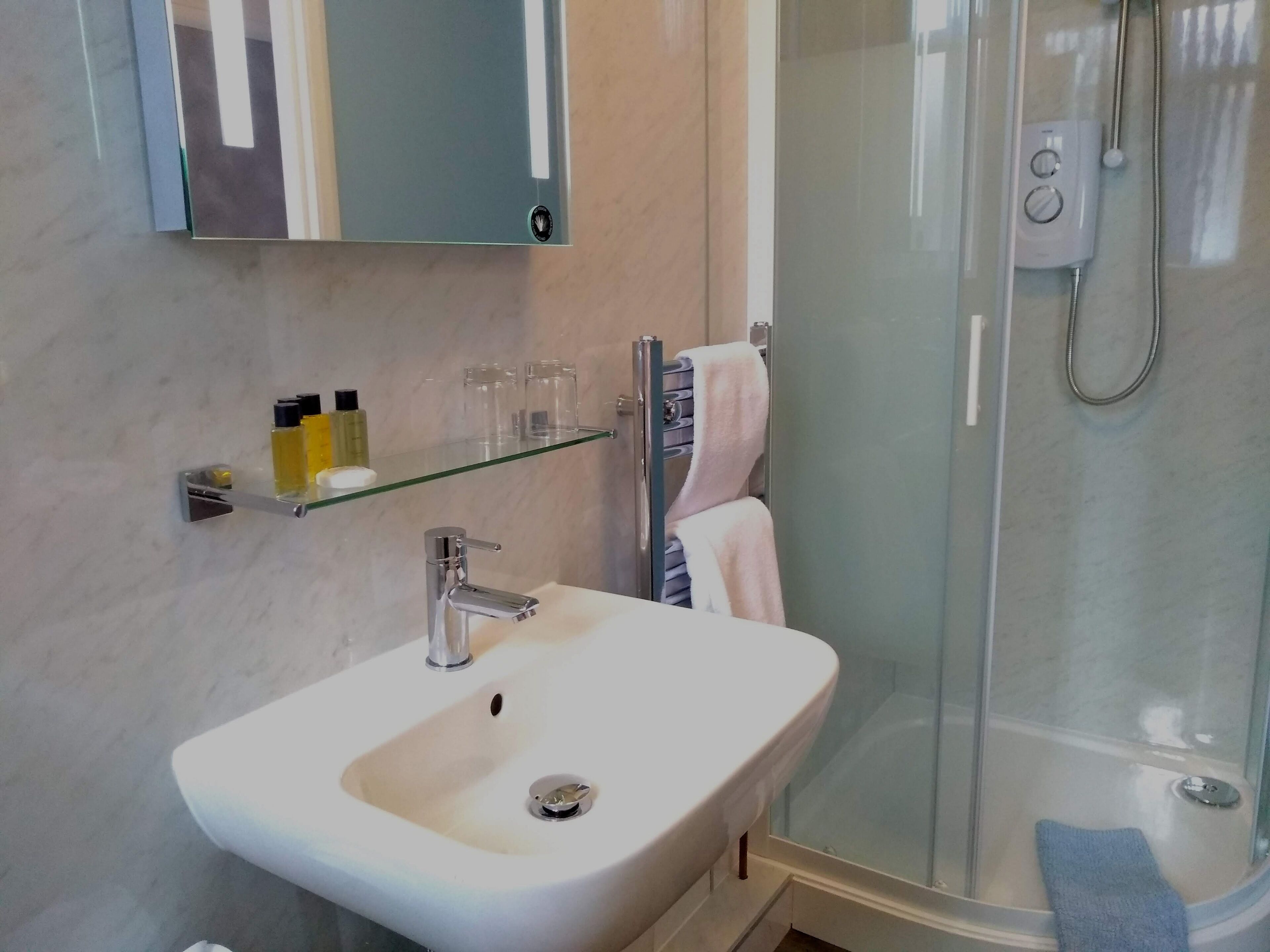 A bathroom with a white sink, glass shelf with toiletries, a towel rack, and a glass-enclosed shower.