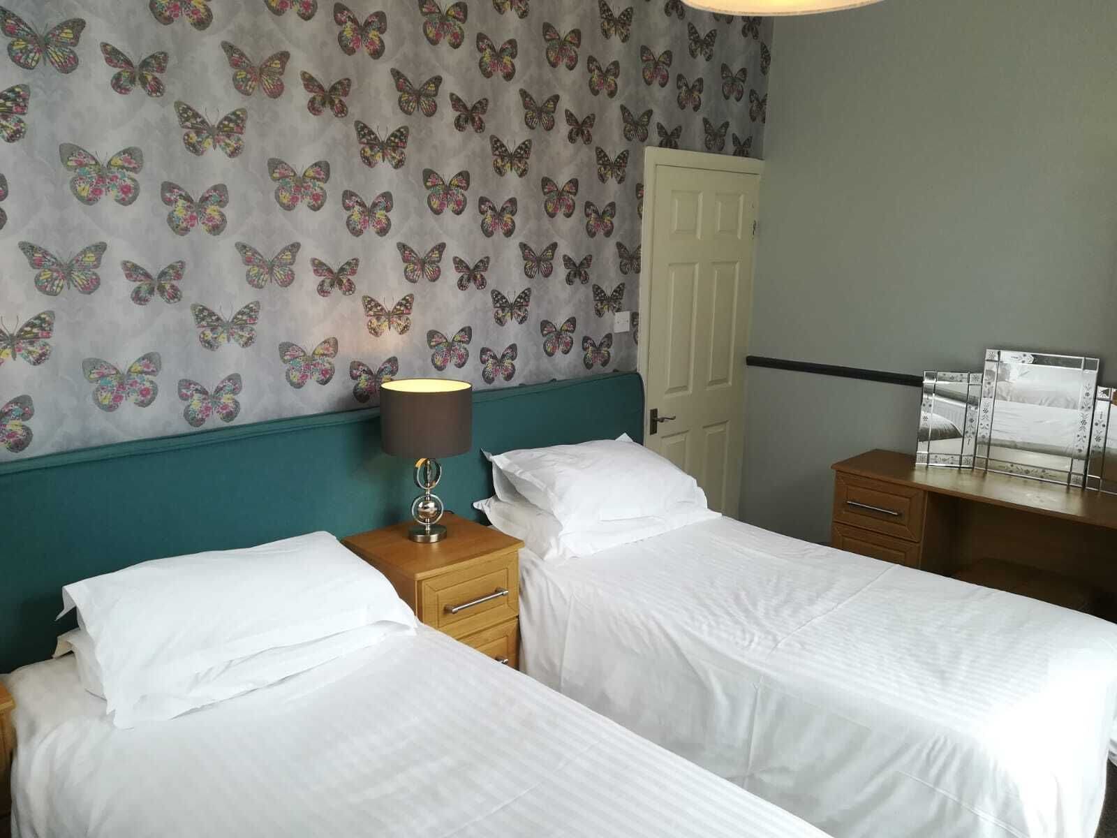 A hotel room with two beds, a nightstand with a lamp, a wooden dresser, and a wall with a butterfly pattern.