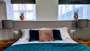 A neatly made bed with a teal duvet, two bedside lamps, and a window with a view of buildings. - Bluebird Lodge and Waterhead Apartment (Coniston)