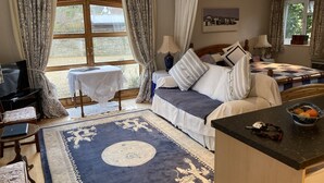 Un dormitorio bien iluminado con cama, mesitas de noche, televisor y vistas al exterior a través de amplias ventanas.
