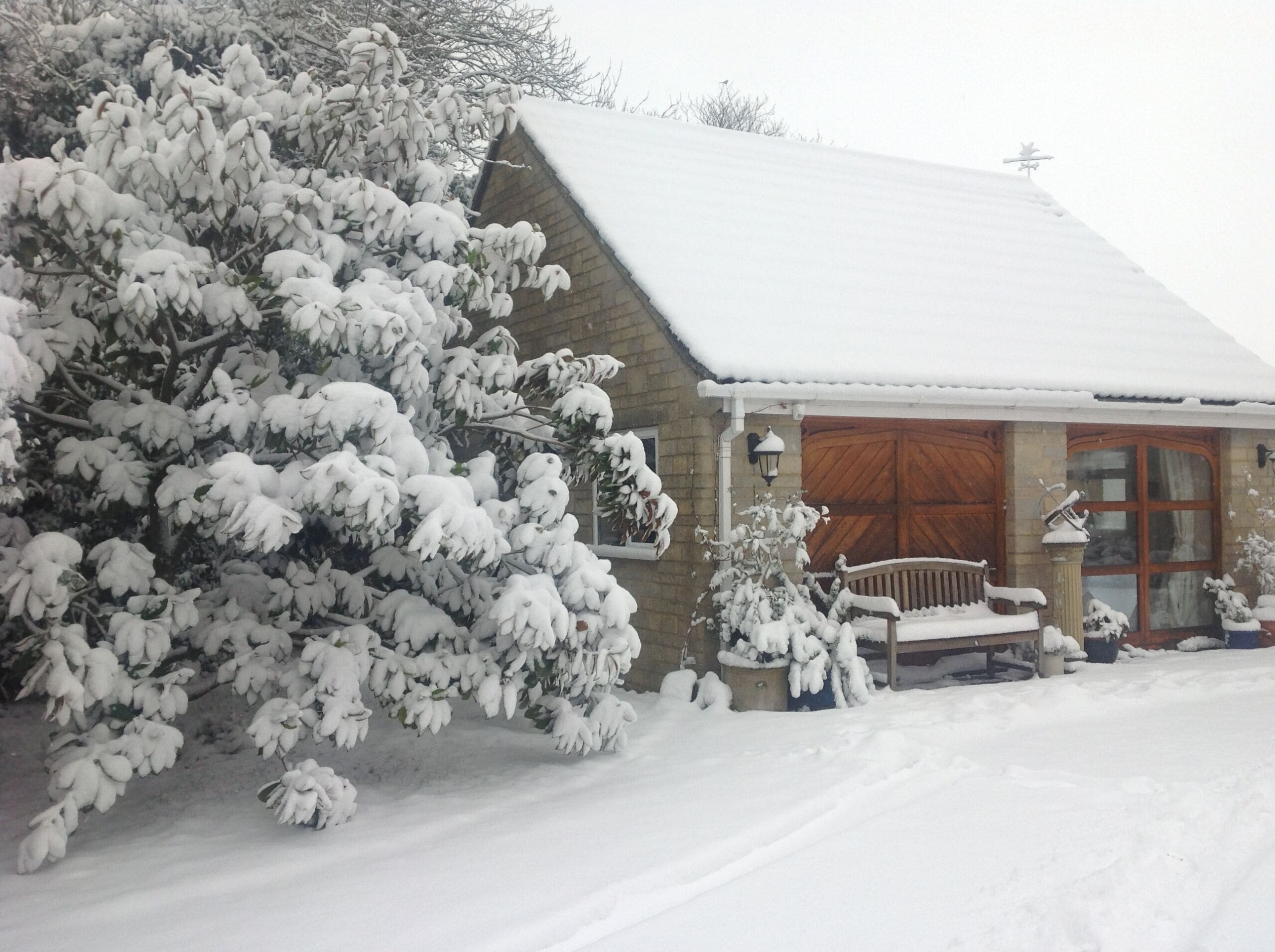 A snow-covered house with a wooden exterior, a large window, and a porch with a bench.