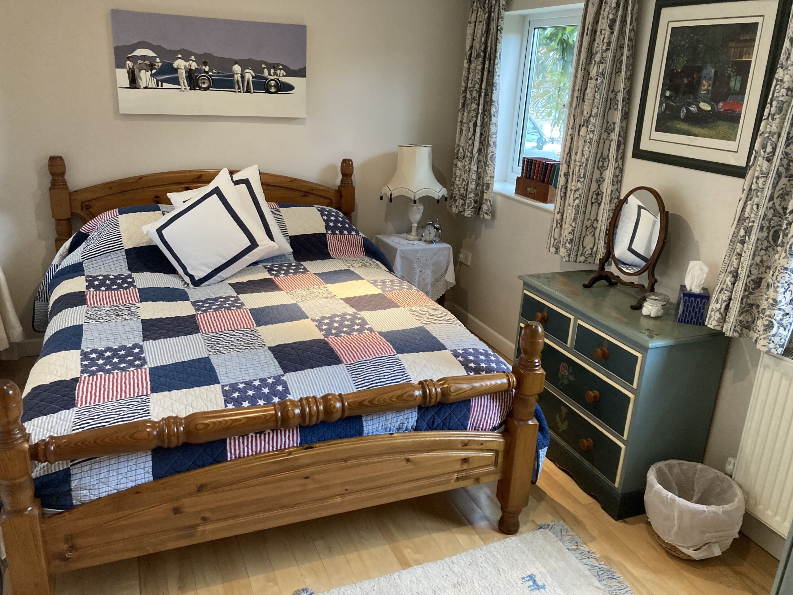 A bedroom with a wooden bed, a quilted bedspread, a dresser, a mirror, a window with curtains, and a wall-mounted picture.