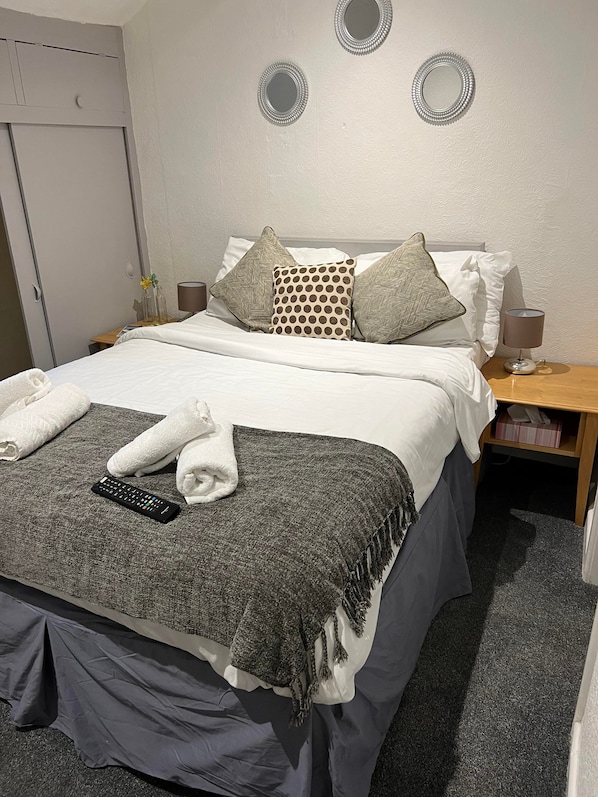 A neatly made bed with a remote control, towels, and a grey throw blanket. There are two bedside tables with lamps and a mirror on the wall. - The Wilton Hotel (Blackpool)