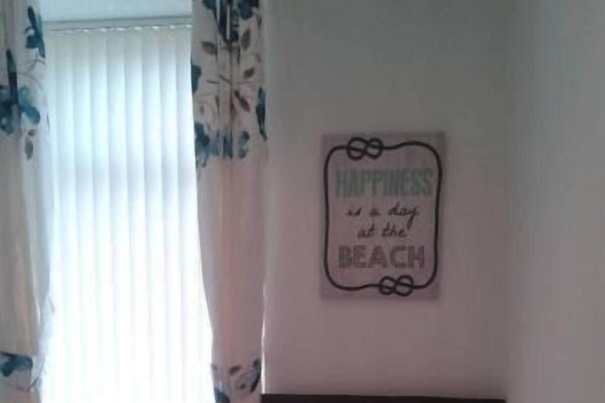 A neatly made bed with a teal bedspread, a pillow, and a remote control on it. There is a wall-mounted sign with the words "Happiness is a day at the beach." A window with sheer curtains is visible, and a small wooden nightstand is next to the bed.