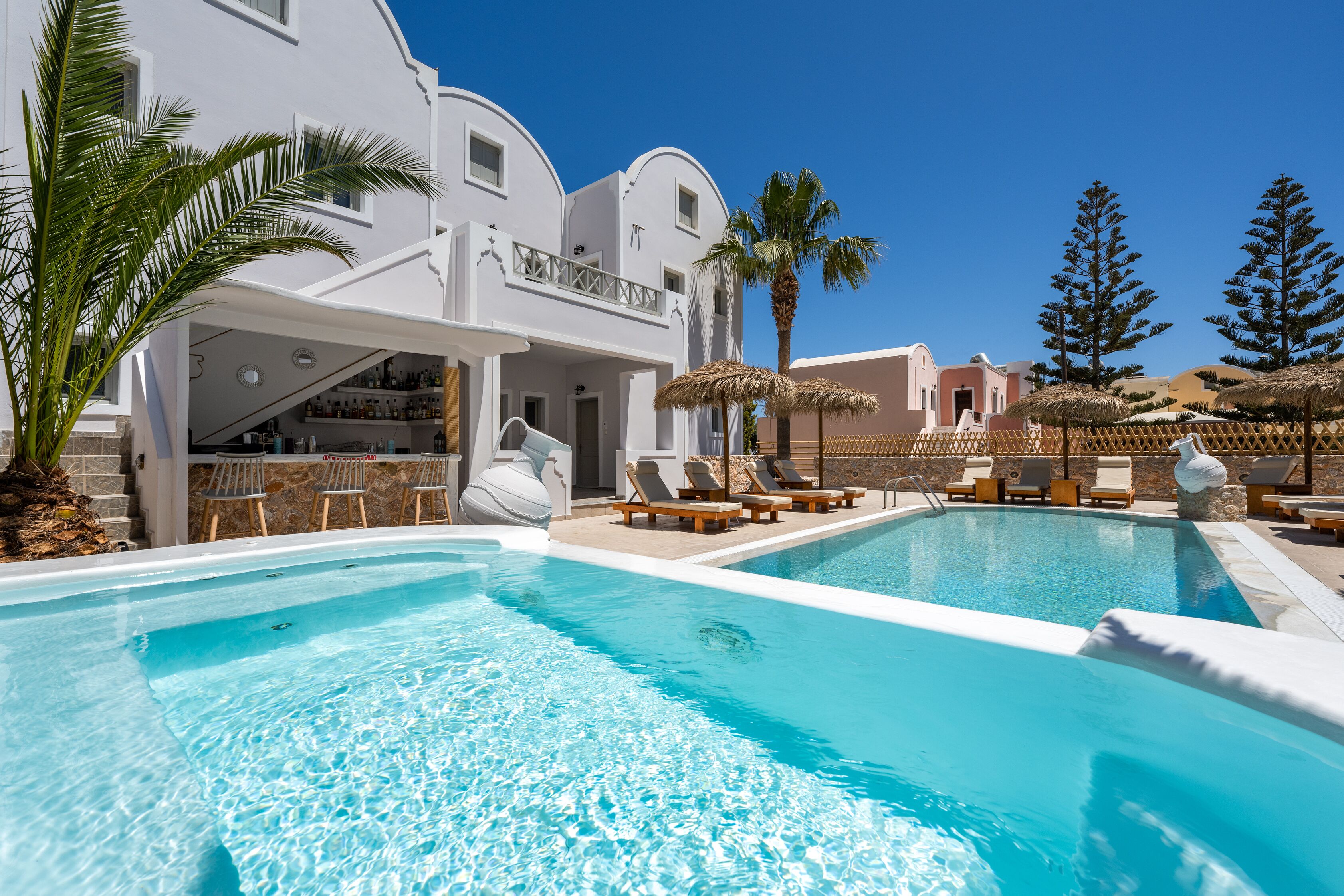 Un área de piscina con sillones de descanso y sombrillas, un bar y un edificio con fachada blanca.